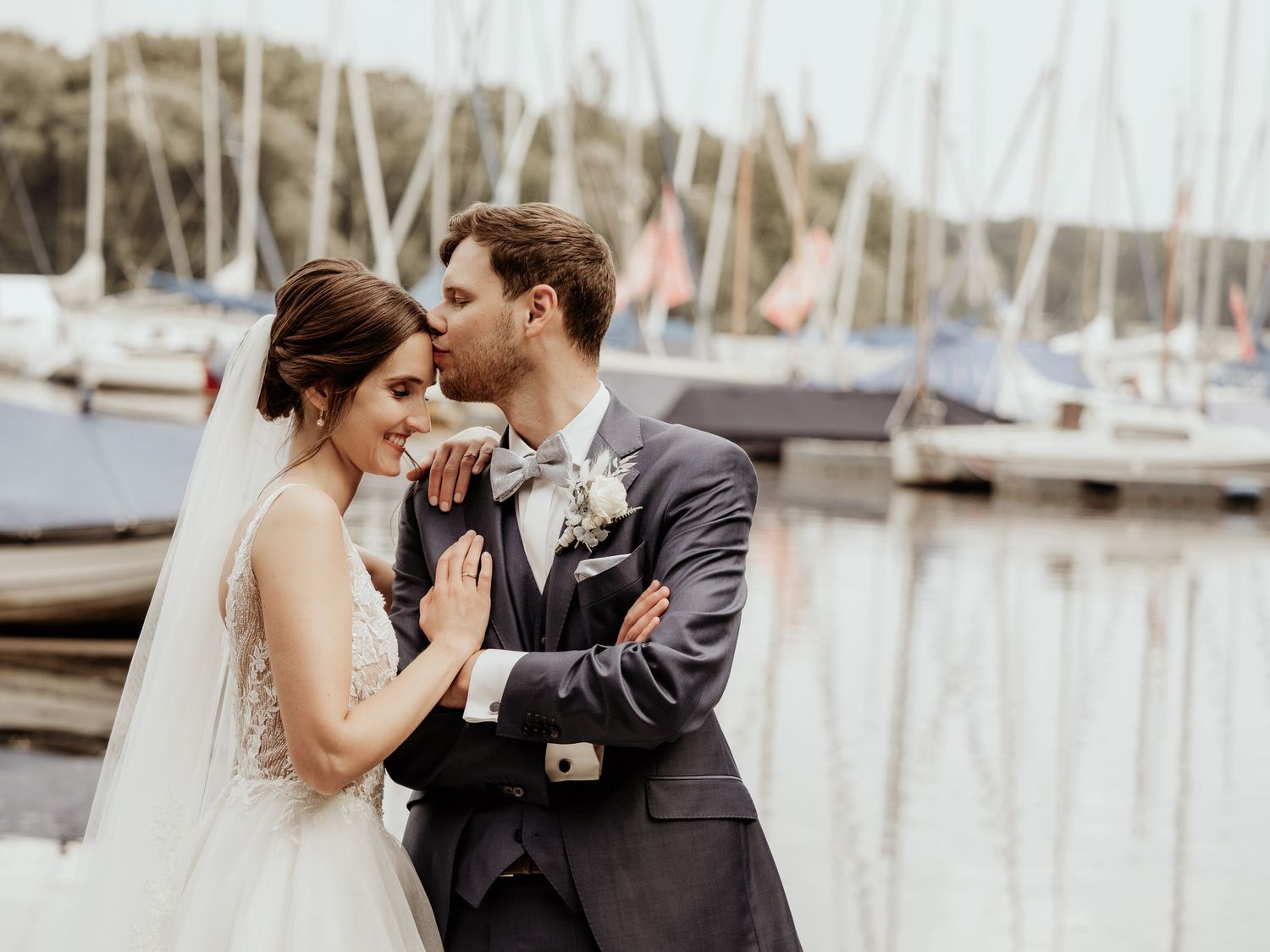 A romantic wedding couple shares an intimate moment by a marina filled with sailboats on a misty day.