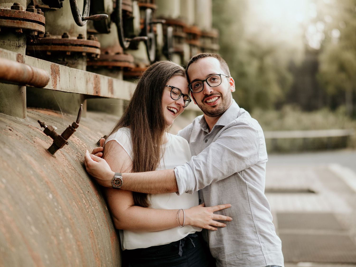 A couple embraces and smiles together while standing next to rustic wooden wine barrels in an outdoor setting.