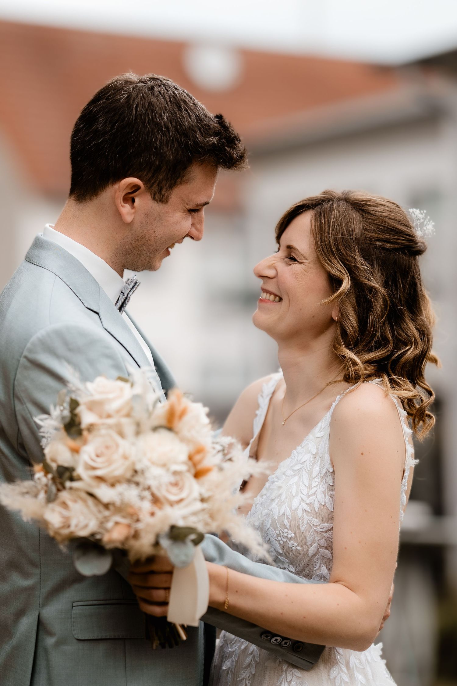 Smiling bride and groom share a tender moment holding a bouquet of blush roses on their wedding day.