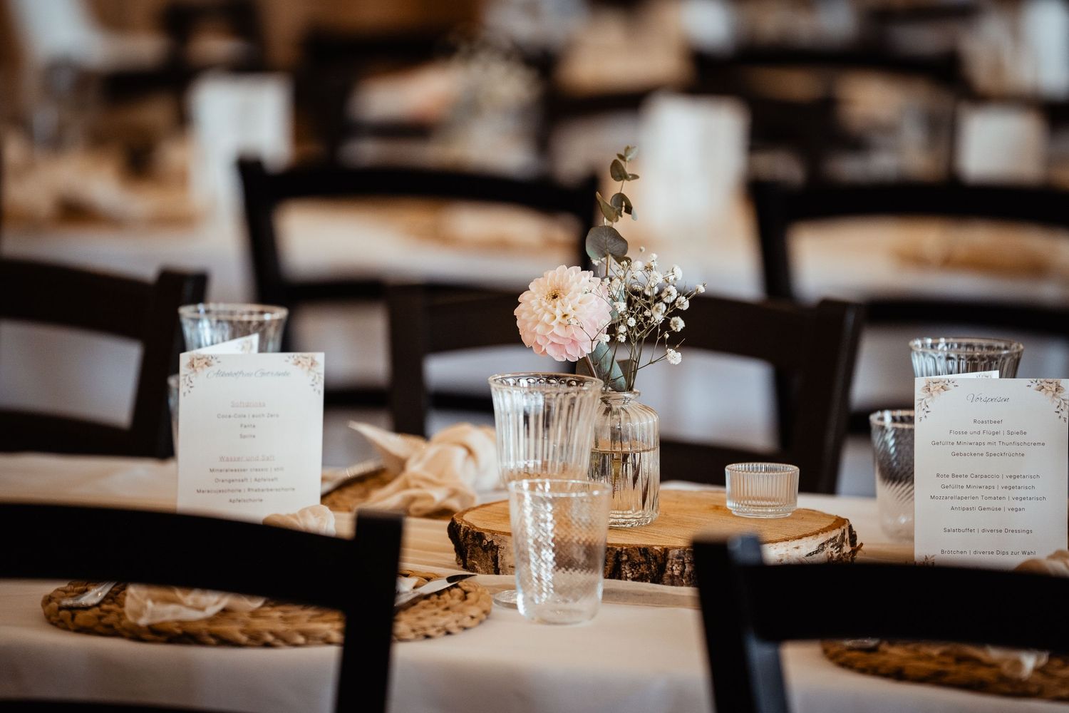Elegant wedding reception table with wood slice chargers, floral centerpiece, menus, and candles on white linen.