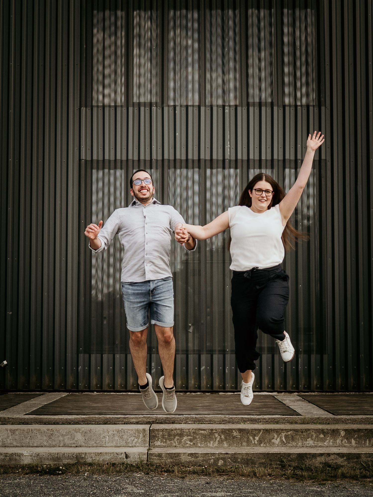 Joyful couple jumping and holding hands in front of a corrugated metal wall, wearing casual summer outfits.
