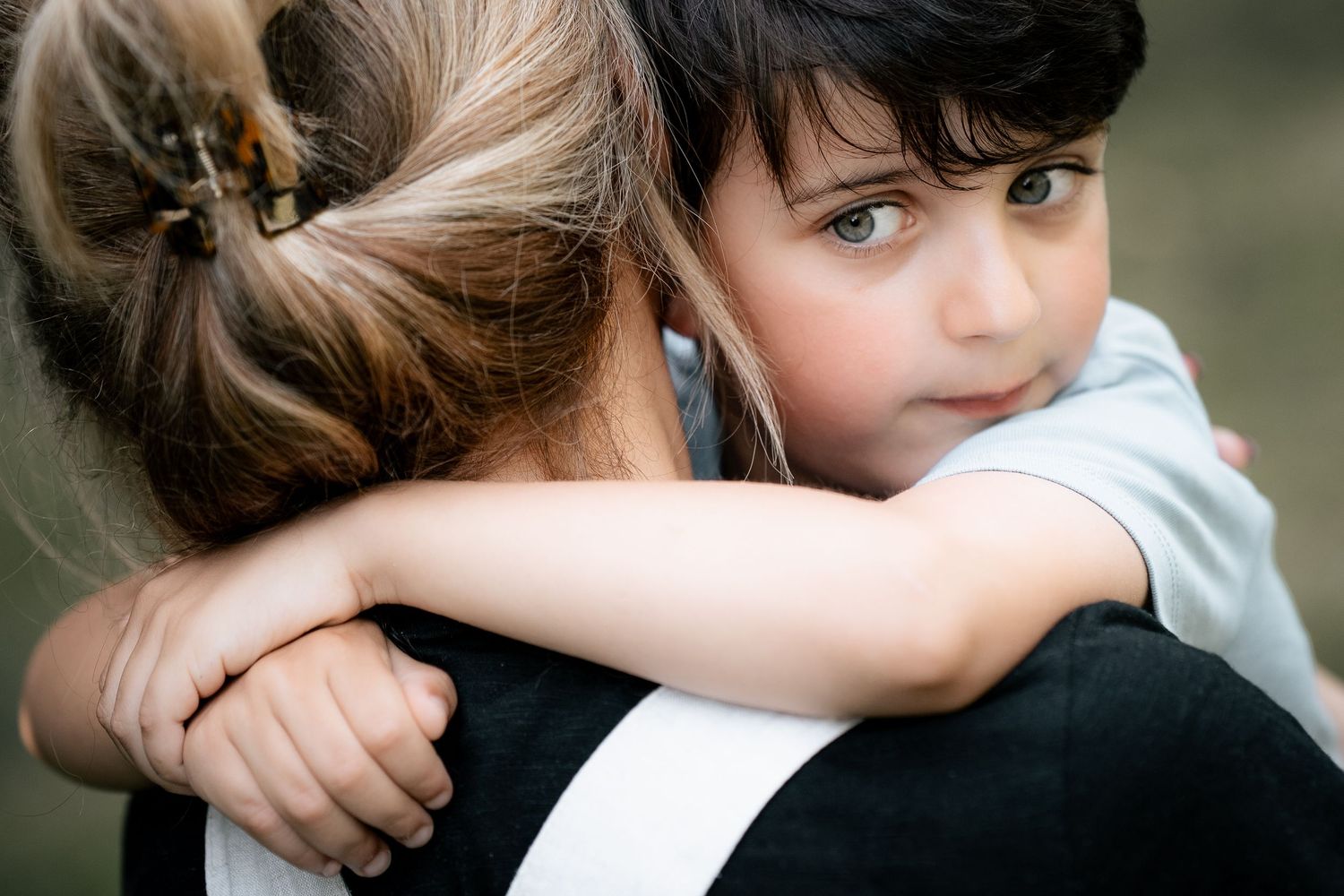 Young child with green eyes hugging an adult, looking over their shoulder in a tender embrace outdoors.