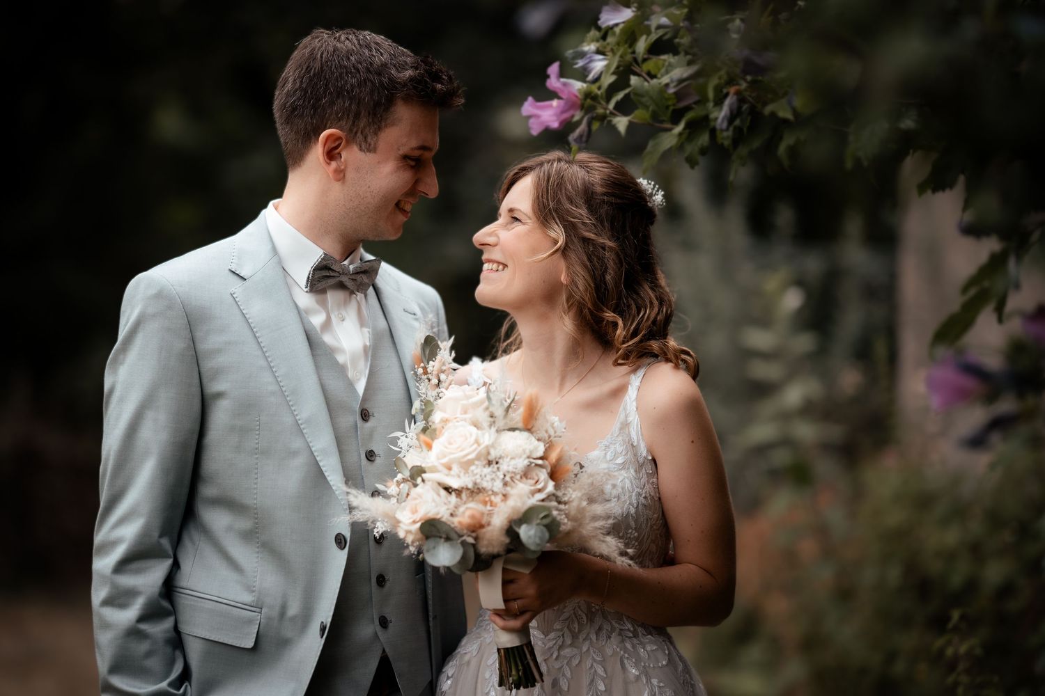 A smiling bride holding a white bouquet stands with a groom in a light blue suit outdoors near blooming flowers.