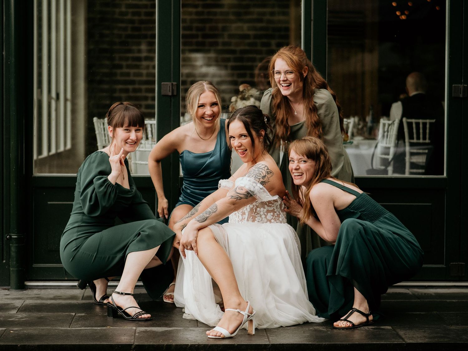 Bride in white gown poses with four bridesmaids in dark green dresses outside a wedding venue.