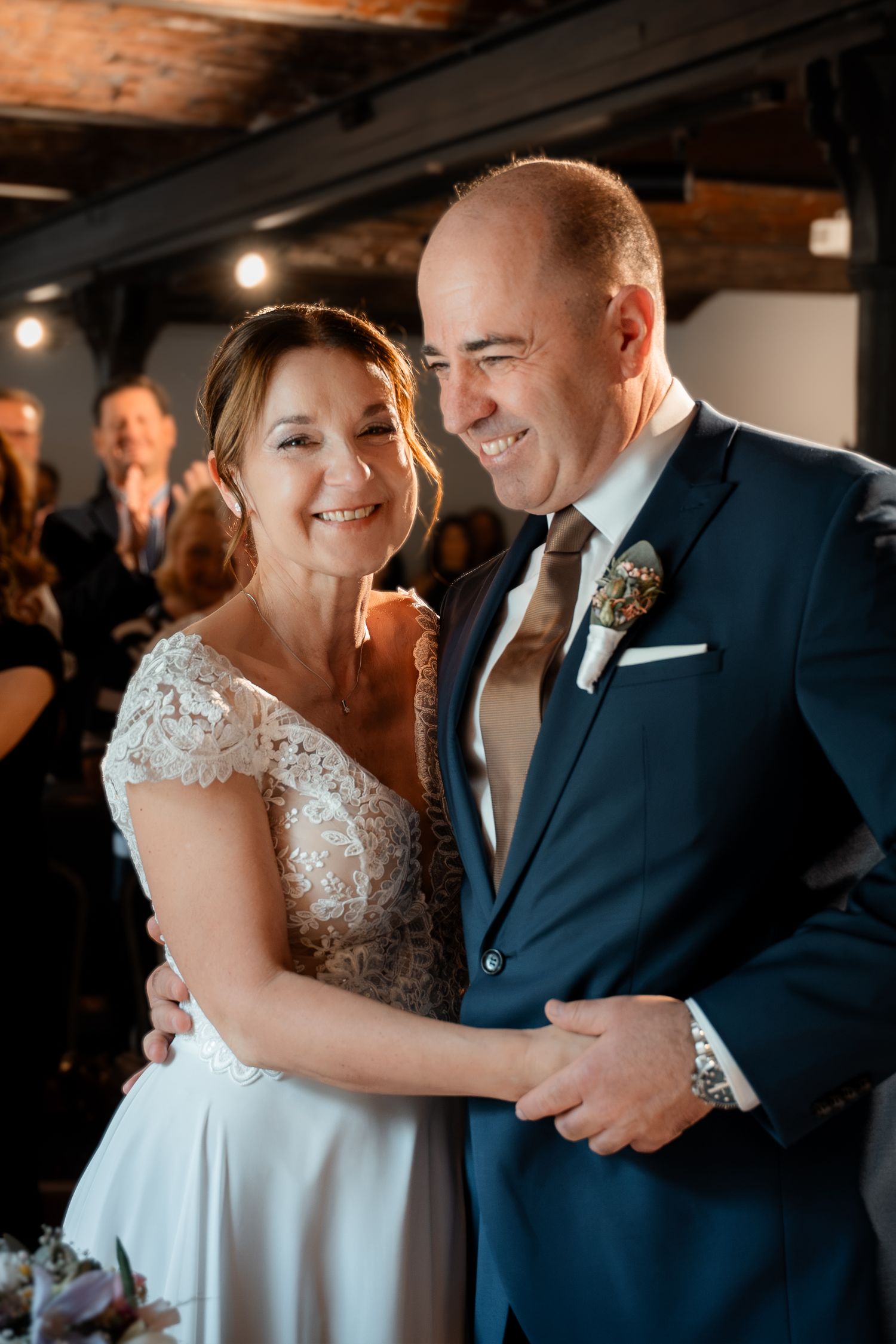 Smiling bride in lace dress and groom in blue suit embrace warmly during indoor wedding reception celebration.