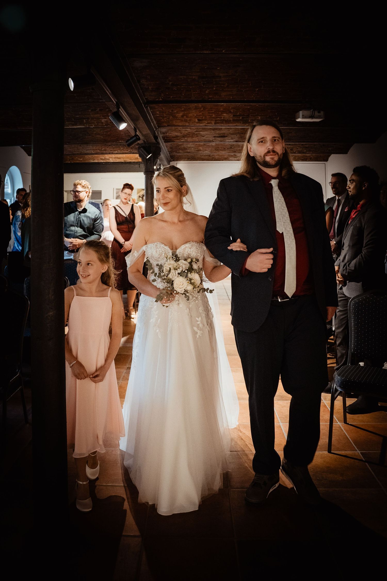 Bride in white gown walks down aisle with groom in navy suit and young flower girl.