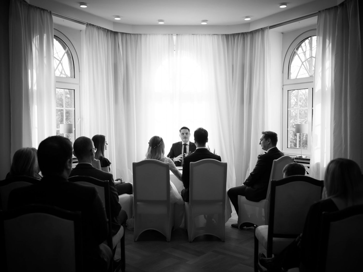 Black and white photo of a small intimate wedding ceremony with guests seated before couple and officiant by bright windows.