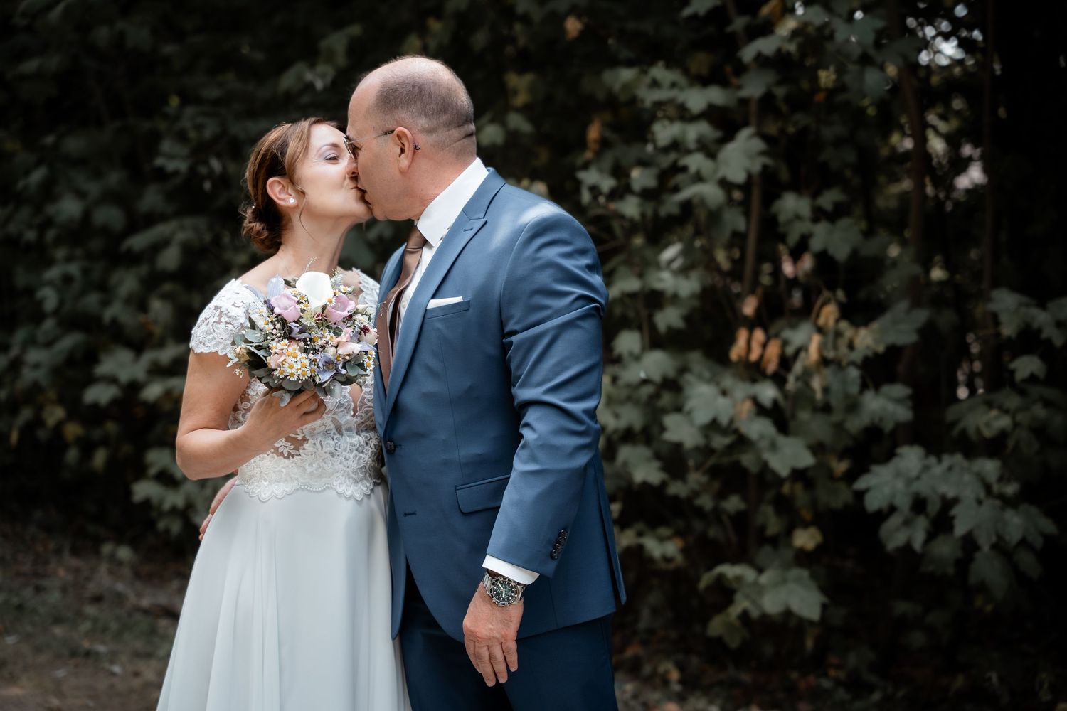 Bride and groom sharing a romantic kiss outdoors, bride holding a bouquet, groom in blue suit.