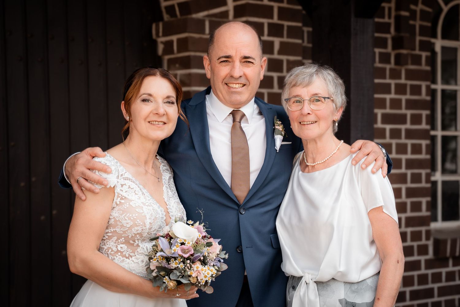 Smiling bride and groom pose with an older woman outside a brick building after their wedding.