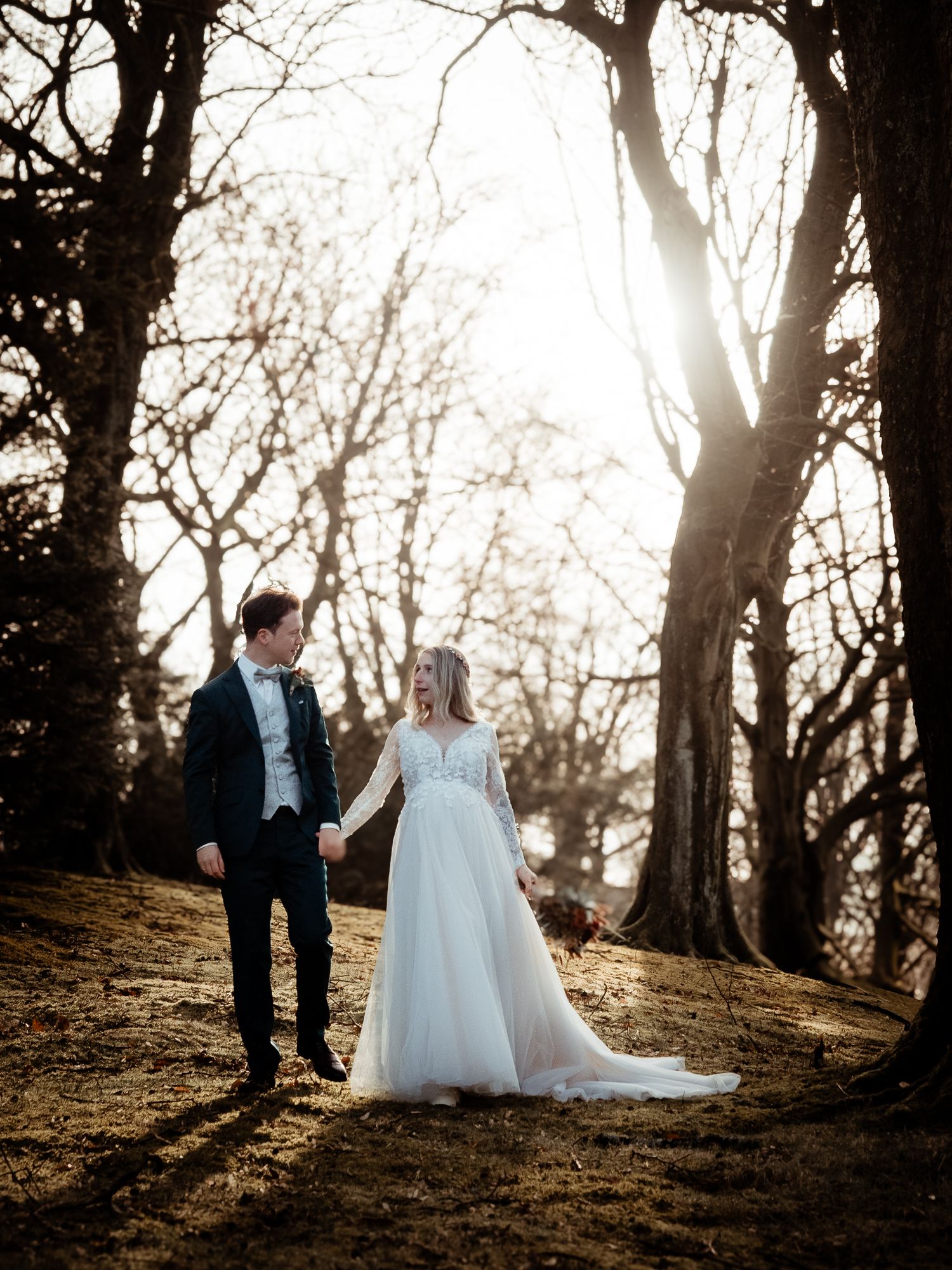 Bride and groom holding hands walking through sunlit winter forest, bride in flowing white tulle gown.