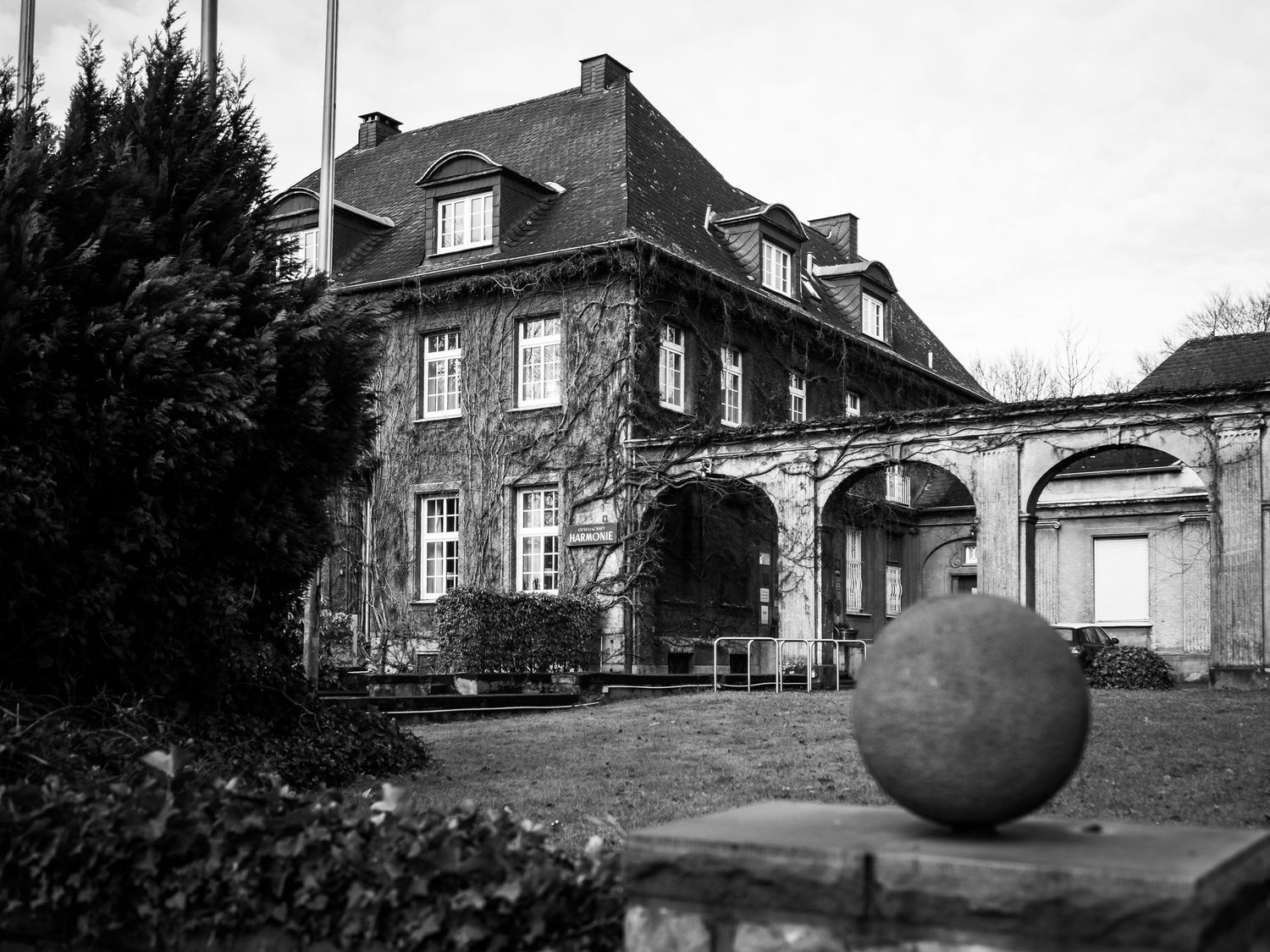 Black and white photo of an ivy-covered historic mansion with a stone sphere sculpture in the foreground.