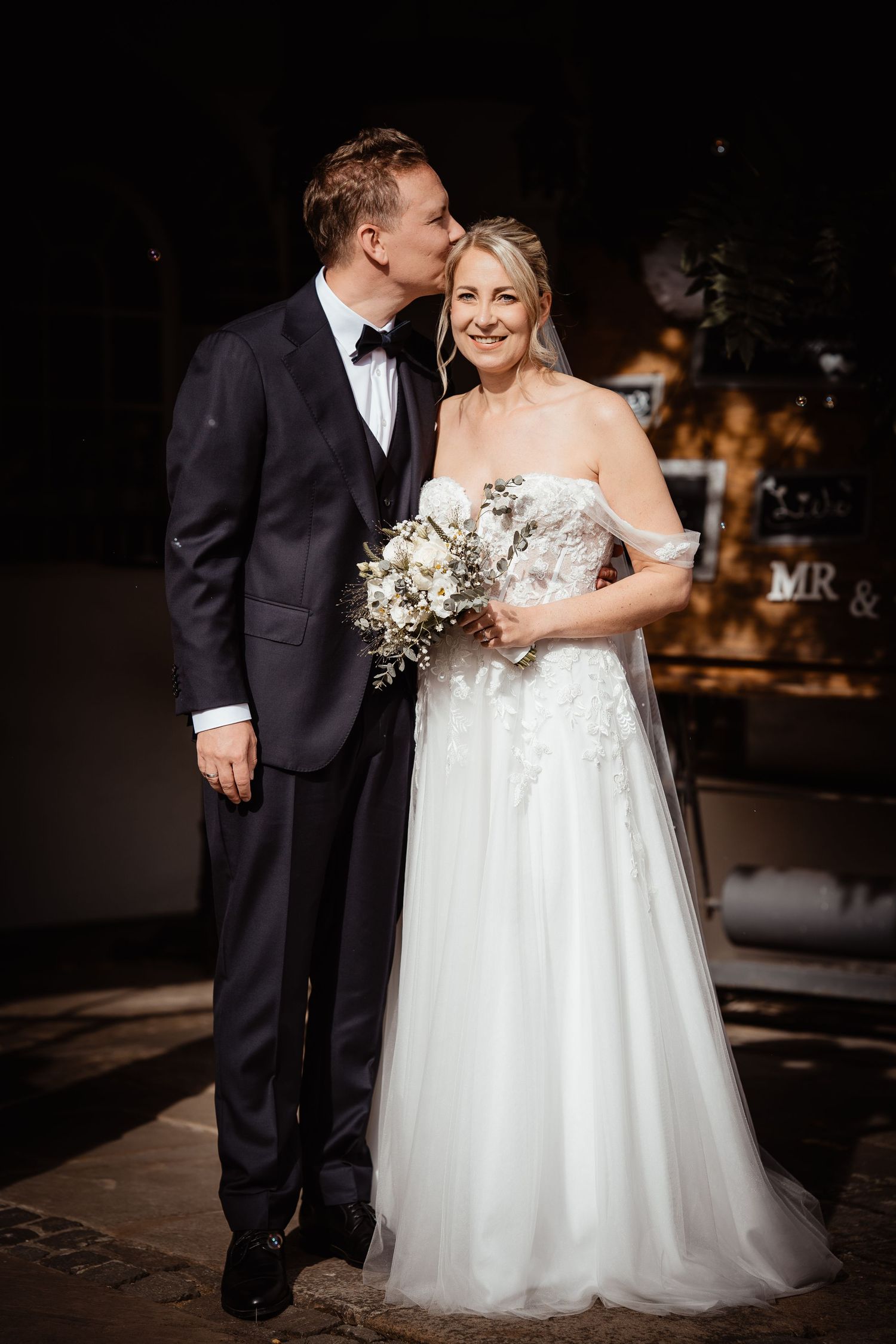 Groom in black tuxedo kissing bride in off-shoulder gown holding white bouquet outdoors.