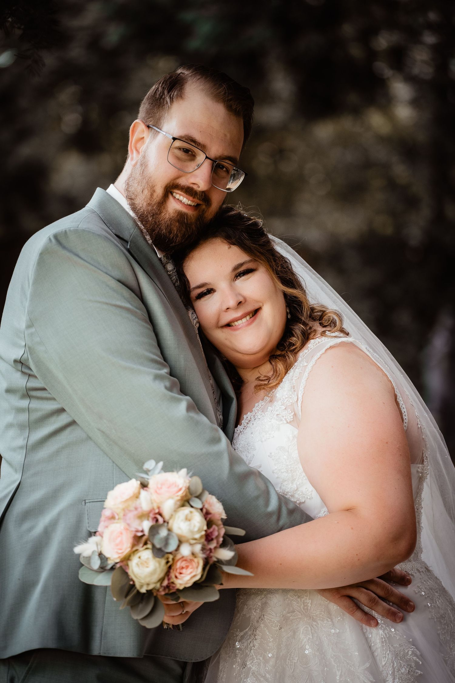 Bride and groom embrace smiling, she holds a pastel bouquet, he wears a gray suit, outdoor wedding portrait.