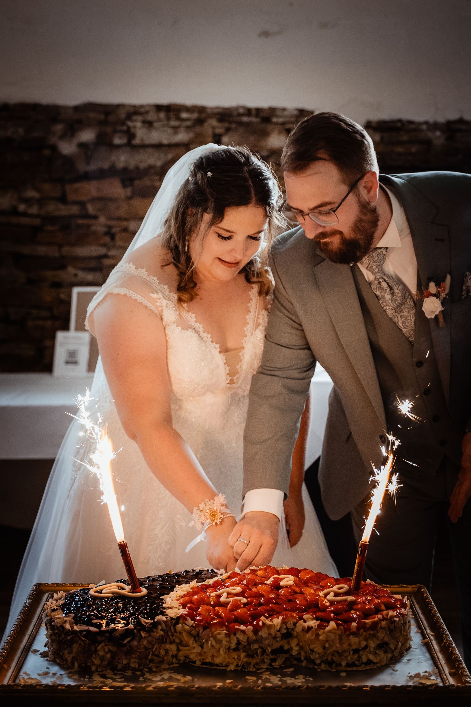 Bride and groom cut a decorative wedding cake with sparklers in a warmly lit rustic reception venue.