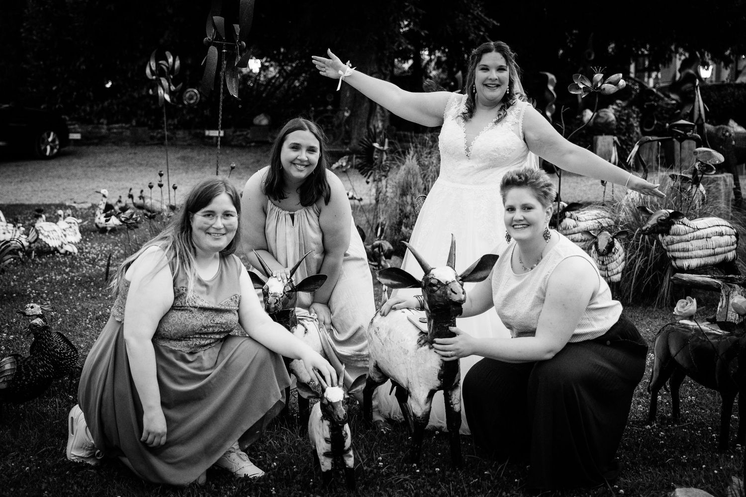 Black and white photo of bride and three bridesmaids posing with goats in decorated garden setting.