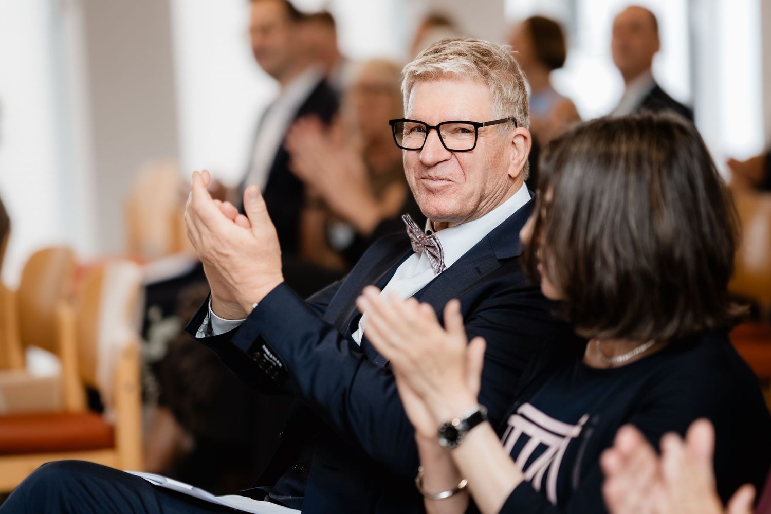 Smiling man in navy suit with glasses applauds at a formal event while woman beside him claps.