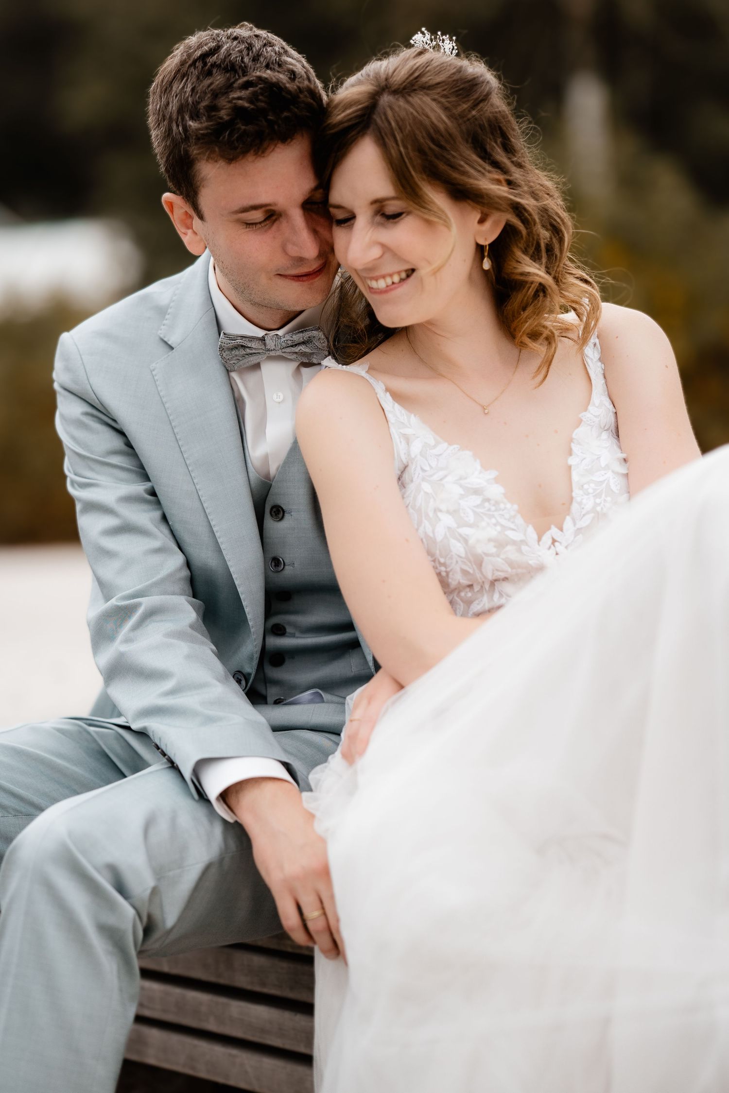 Bride and groom sit closely together on a bench, groom nuzzling bride in a tender wedding portrait.