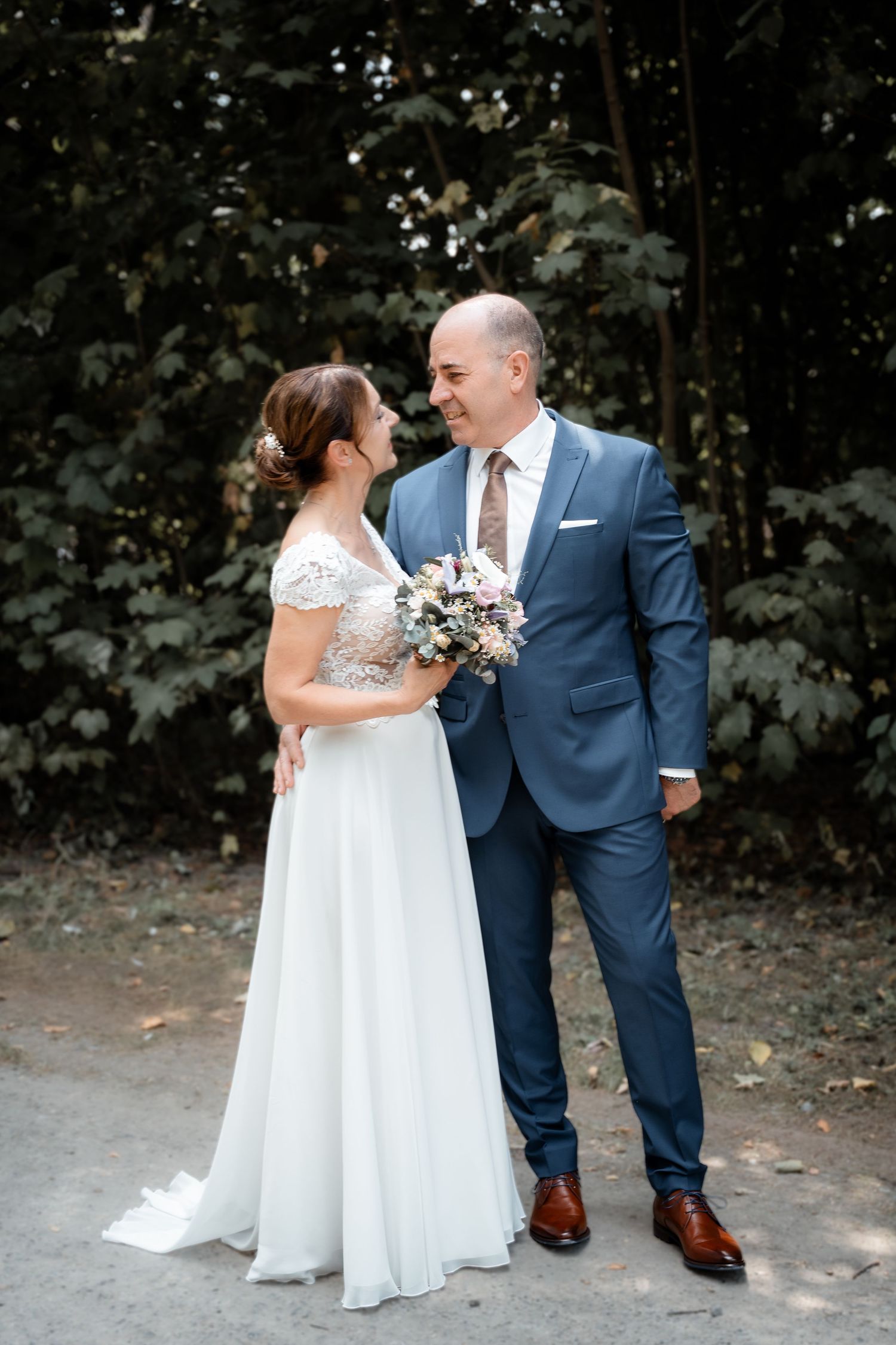 Bride and groom smiling at each other outdoors, she holds a bouquet, he wears a blue suit, green trees behind.
