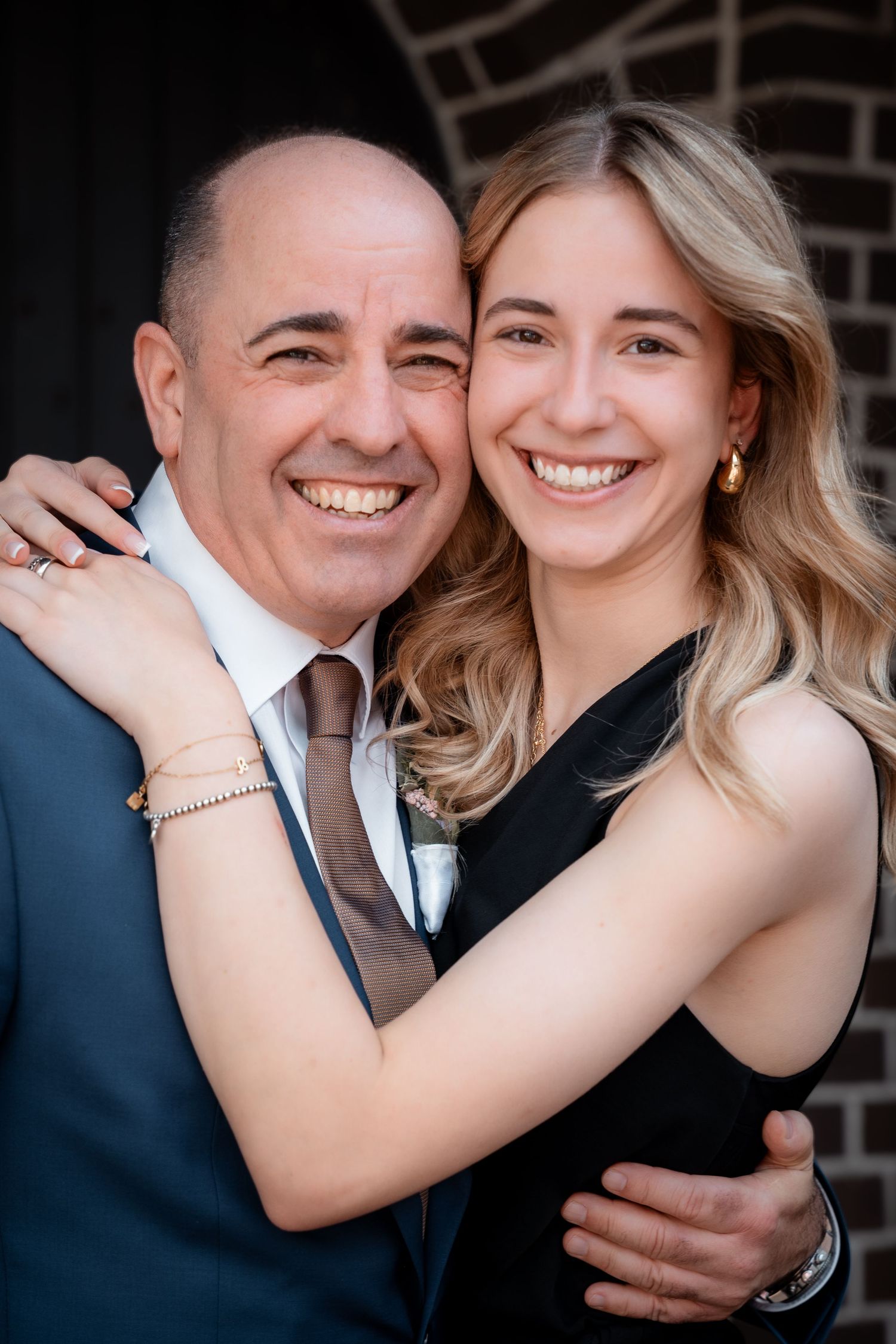 Smiling bald man in suit embraced by young blonde woman in black dress in joyful wedding portrait.
