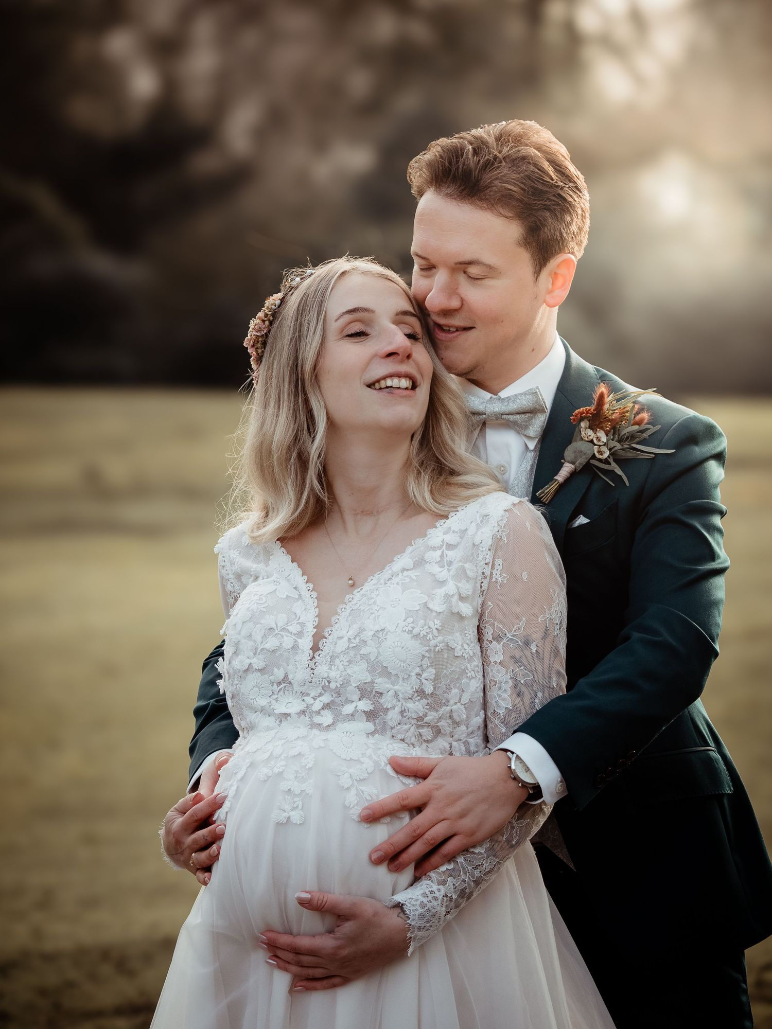 Groom tenderly embraces pregnant bride in lace wedding dress during golden hour outdoor wedding portrait.