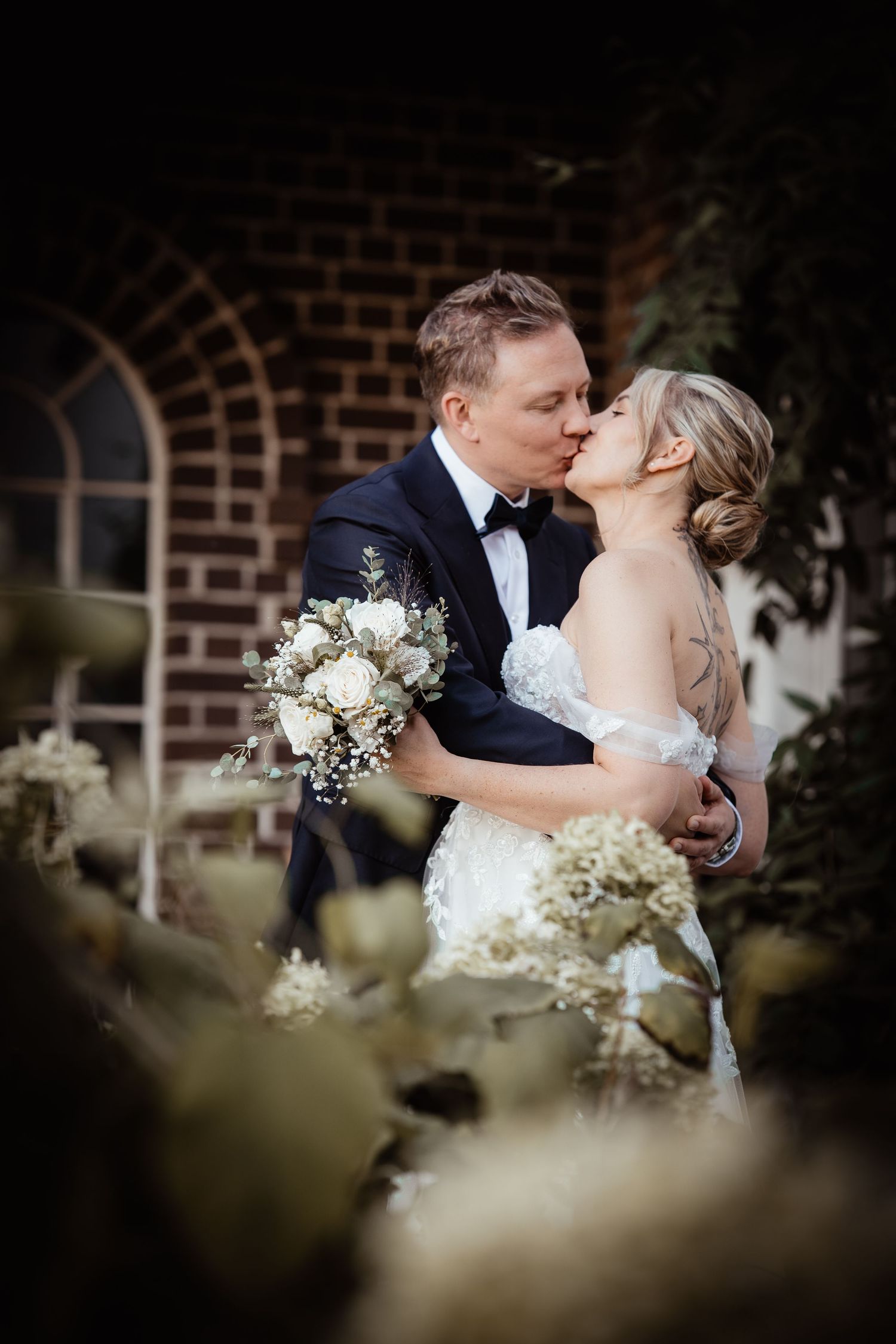 Bride and groom sharing a romantic kiss while holding a white bouquet in front of a brick wall.