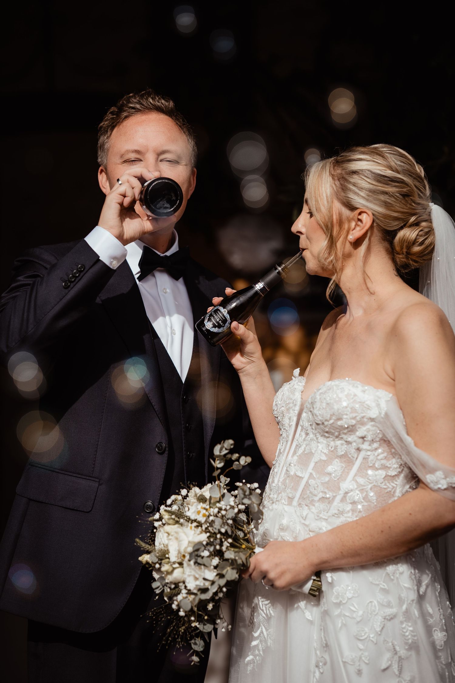 Bride and groom in elegant attire share a toast drinking from beer bottles at wedding.