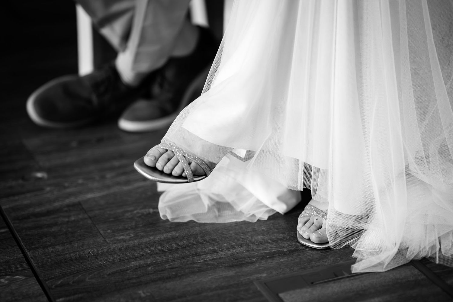 Black and white photo of bride in white gown wearing sandals beside groom in sneakers on wooden floor.