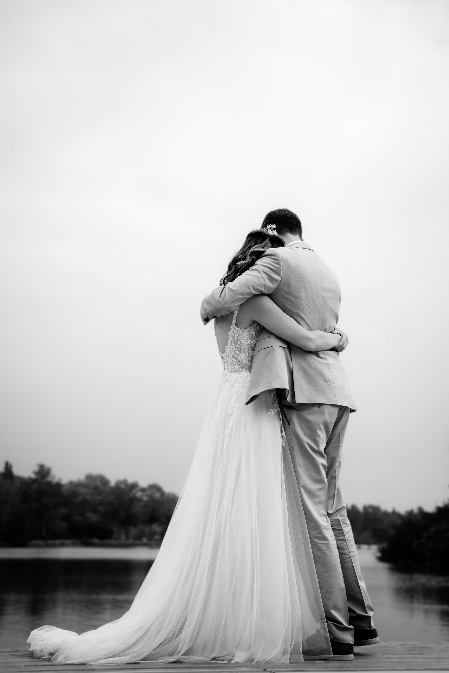 Bride and groom embracing on a dock by a lake in a romantic black and white wedding photo.