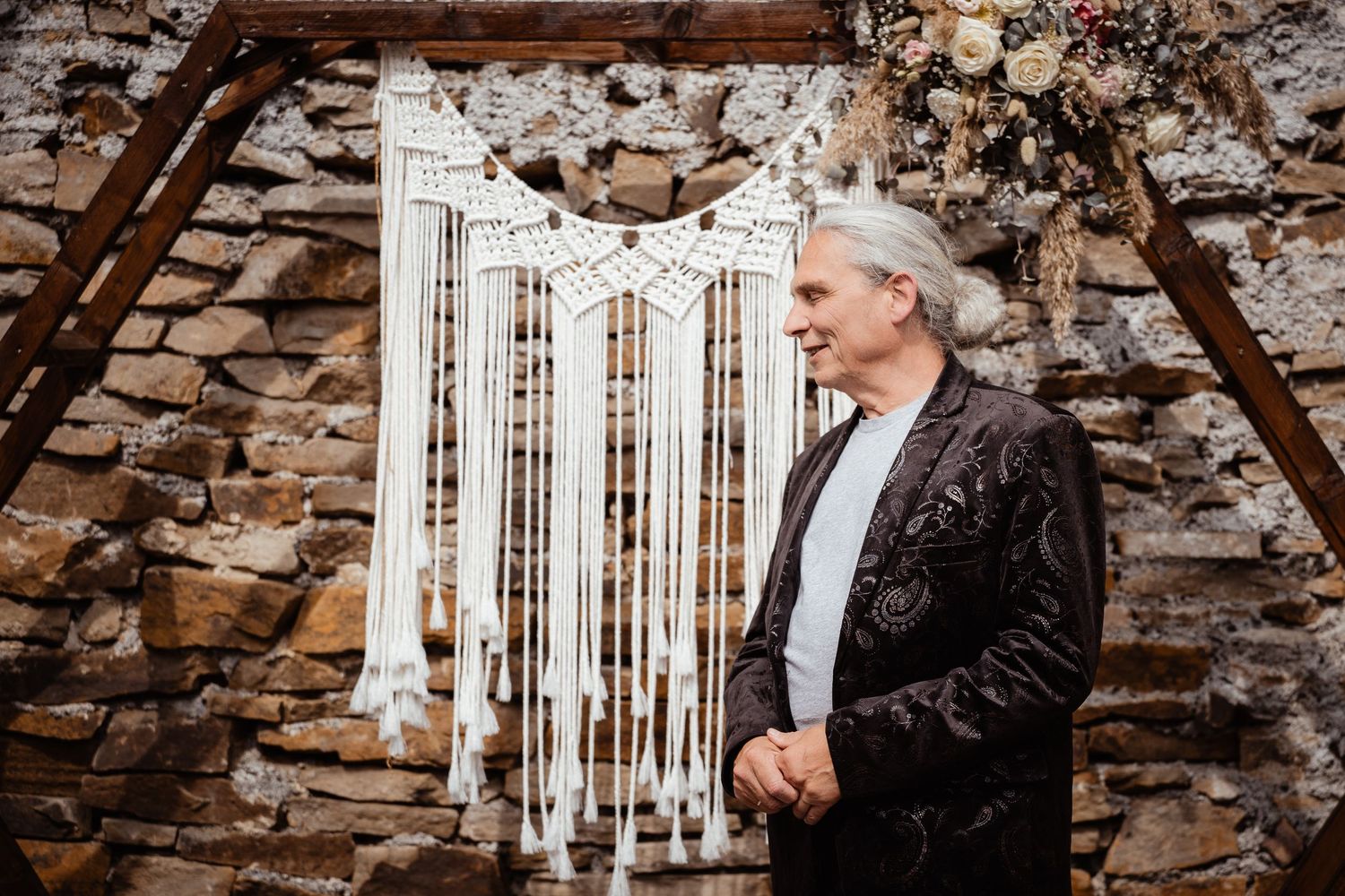 Older man in dark velvet blazer standing before a macrame backdrop and stone wall at a boho wedding ceremony.