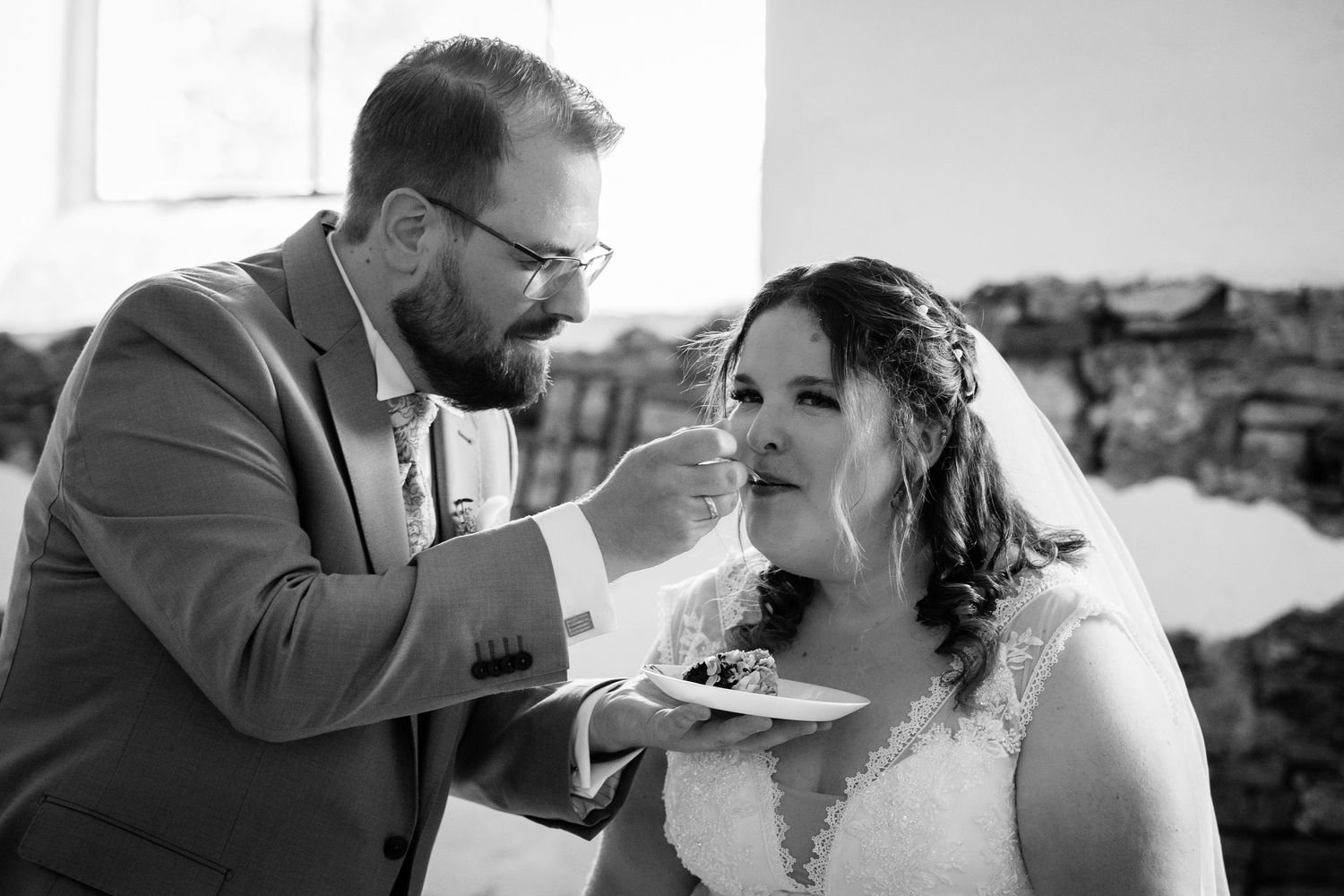 Black and white photo of groom feeding wedding cake to bride wearing lace veil at reception.
