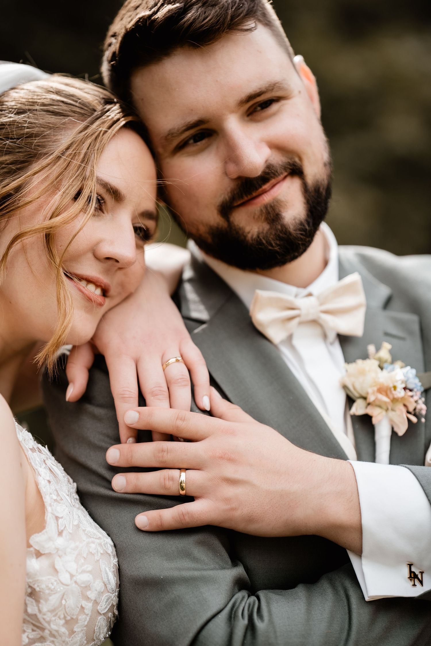 Newlywed couple embracing and displaying wedding rings, groom in gray suit with bow tie and boutonniere.