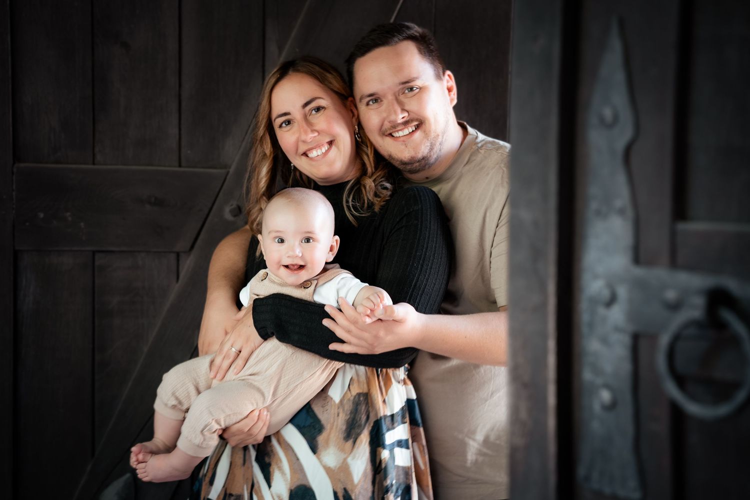 Happy young family posing with smiling baby in front of rustic dark wooden door in professional portrait.