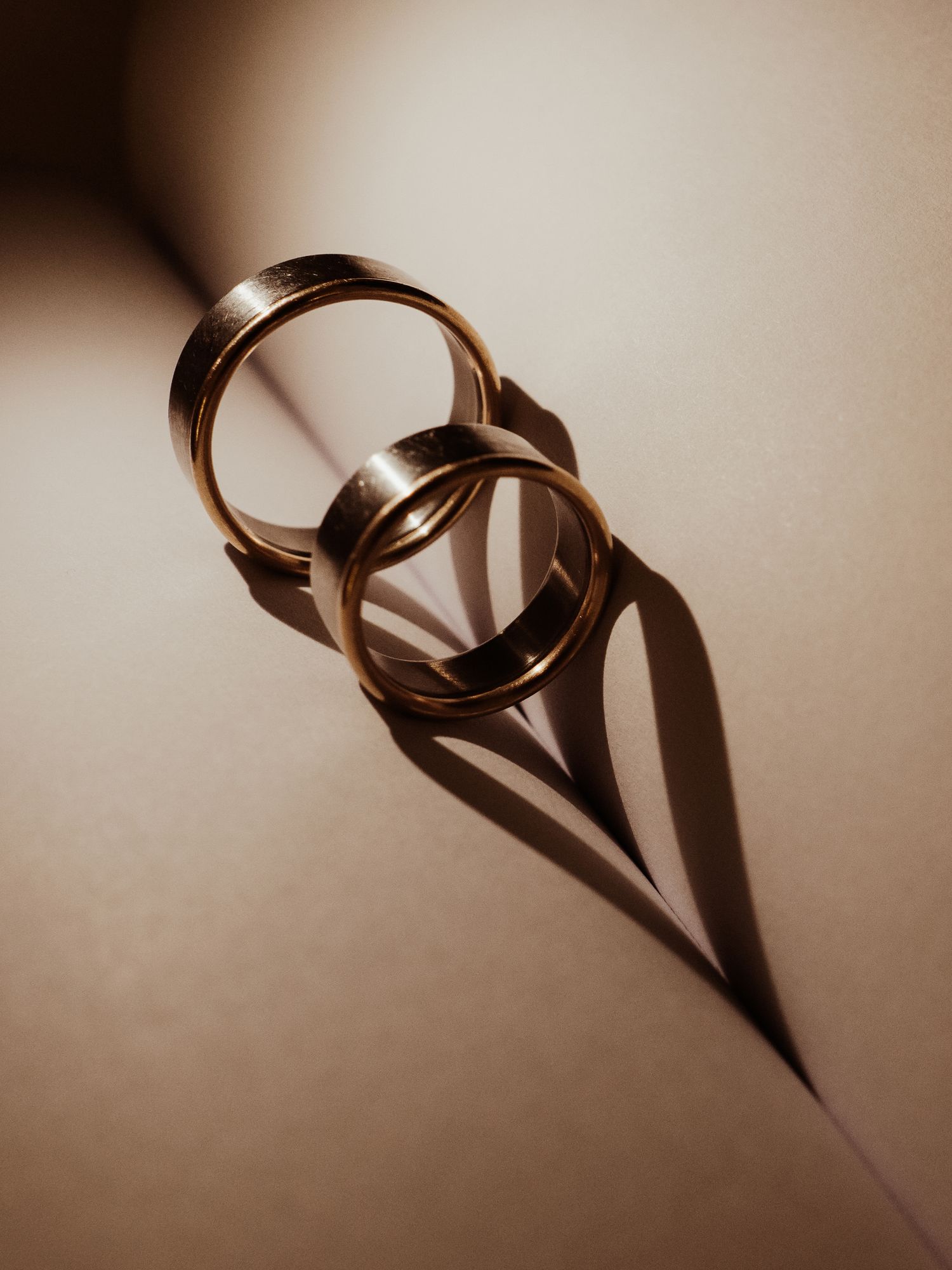 Two gold wedding rings leaning together casting a heart-shaped shadow on a beige surface.