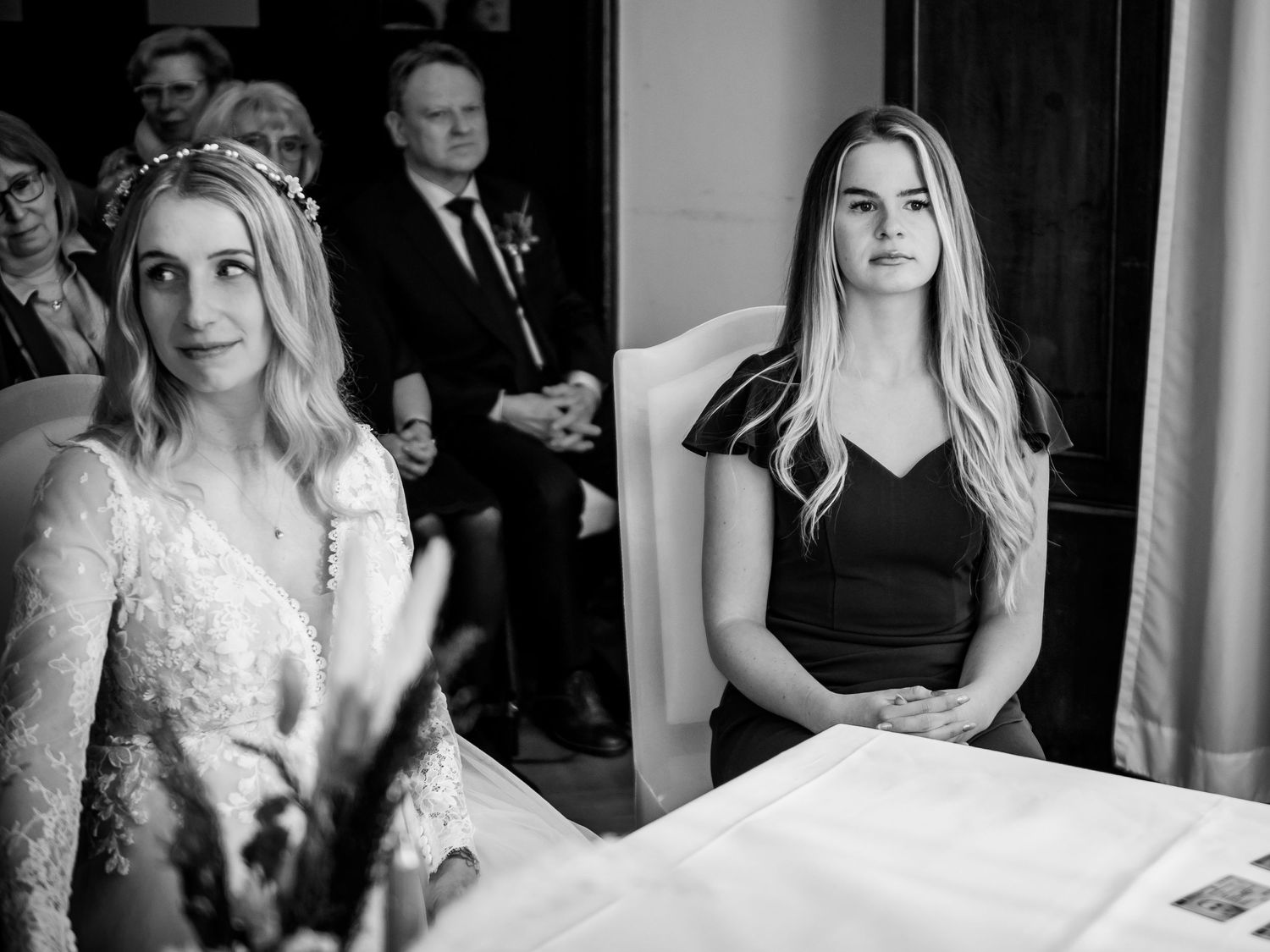Black and white wedding photo of bride in lace gown and bridesmaid in black dress seated at ceremony table.