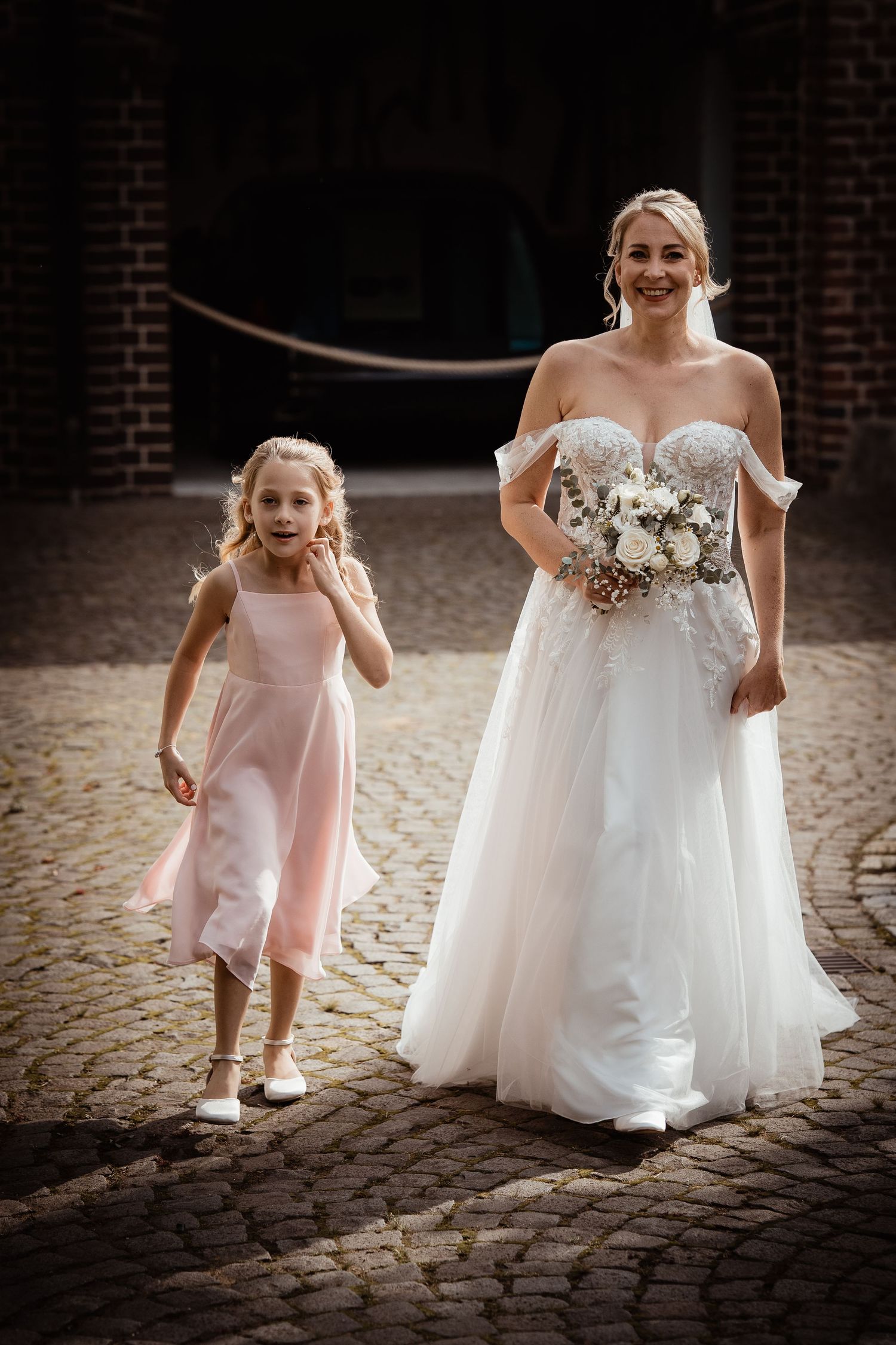 Smiling bride in white off-shoulder gown walks with flower girl in pink dress on cobblestone path.