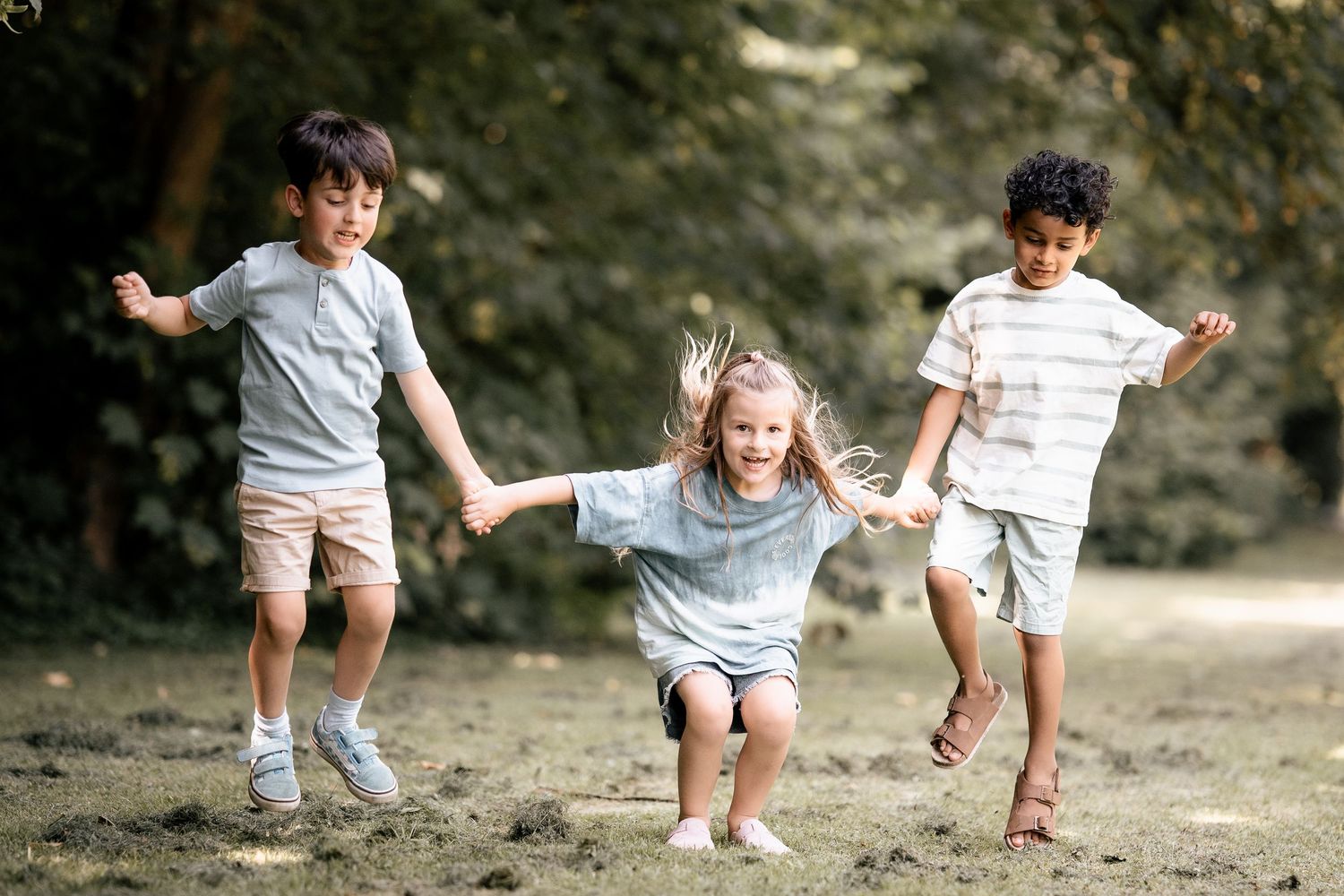 Three happy children holding hands and running together outdoors in a park on a sunny day.