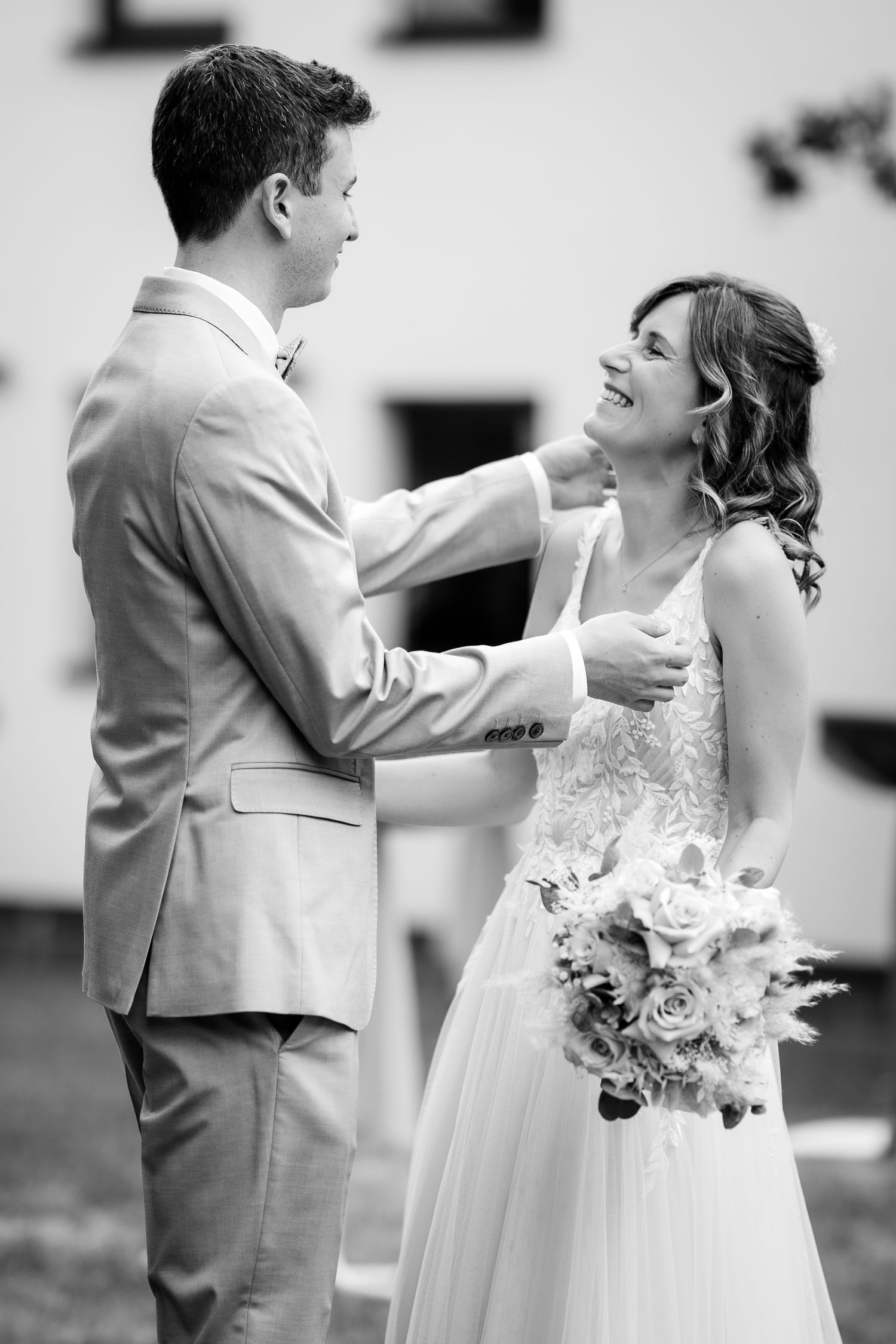A groom in a gray suit tenderly touches his brides face as she smiles holding a bouquet.