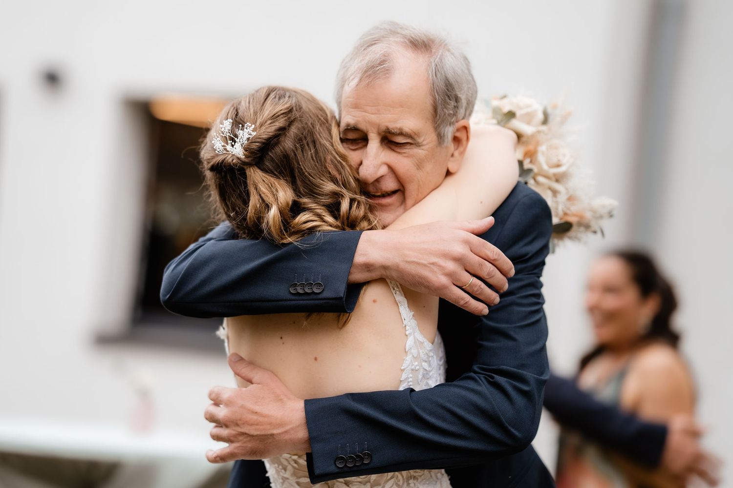 Emotional father-daughter wedding hug, bride in white lace dress with hair accessory embracing older man in navy suit.