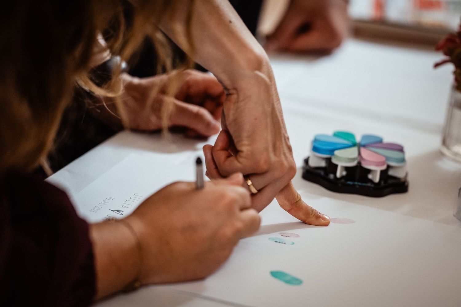 Close-up of hands signing documents at a table with colorful ink pads during a wedding calligraphy activity.