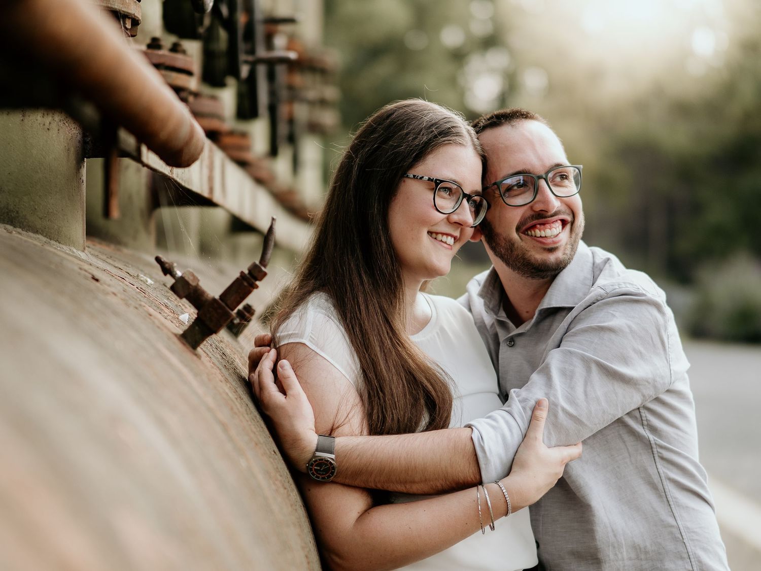 A smiling couple in glasses embrace outdoors, leaning against a rustic metal structure in warm natural light.