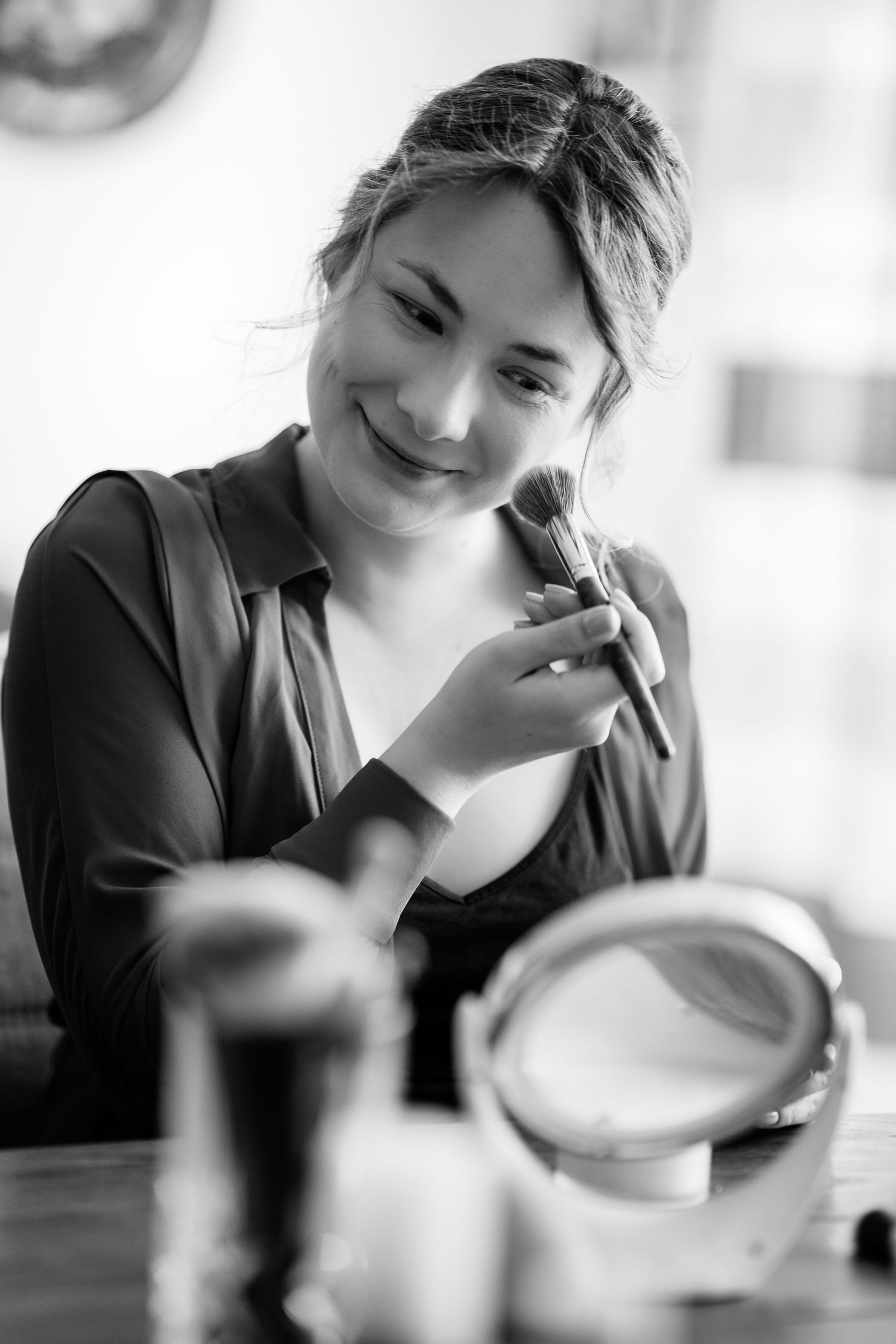 Black and white photo of a smiling woman applying makeup with a brush, looking into a small mirror on a table.