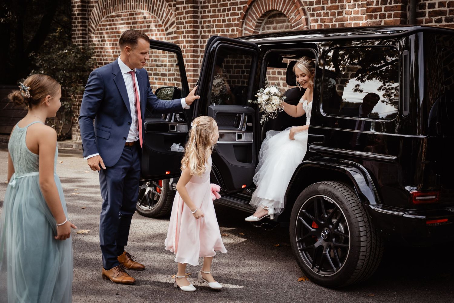 Bride stepping out of black Mercedes G-Wagon SUV at wedding, greeted by groom and flower girls outside brick building.