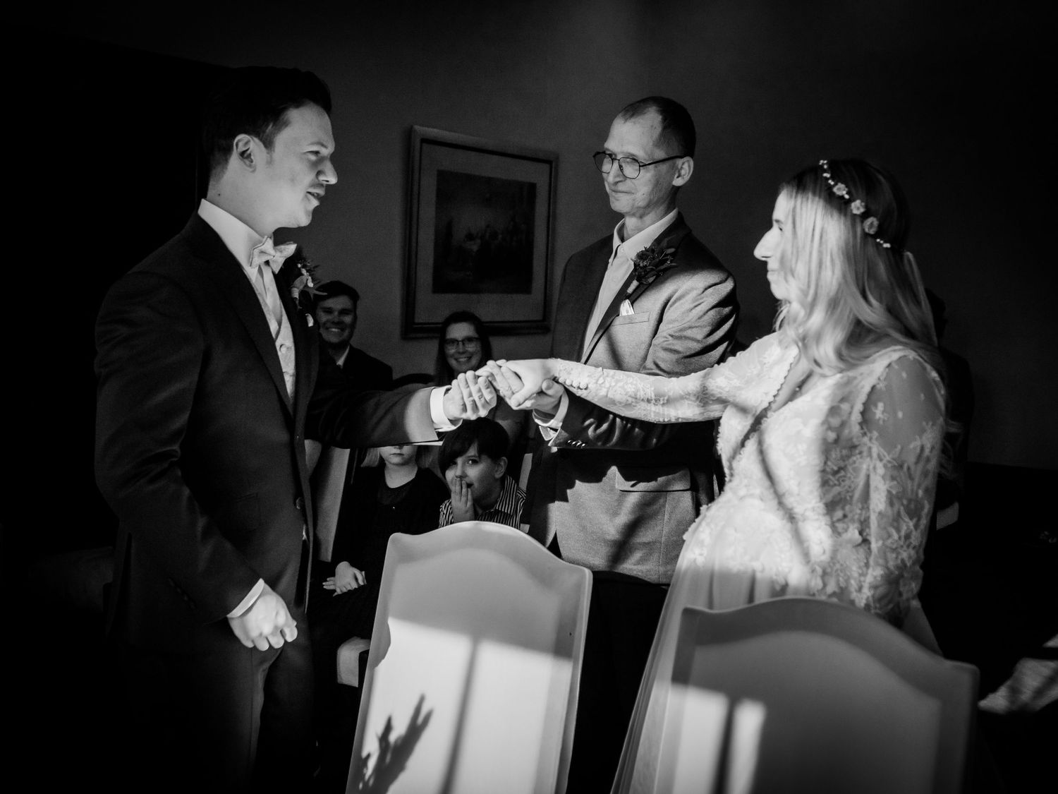 Black and white wedding ceremony photo of couple exchanging rings with officiant and guests in elegant venue.