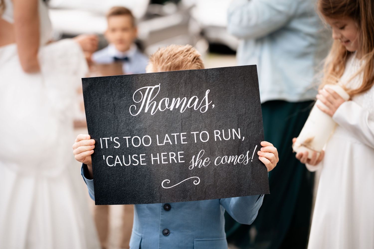 Ring bearer holds funny wedding sign reading Thomas its too late to run cause here she comes at ceremony.