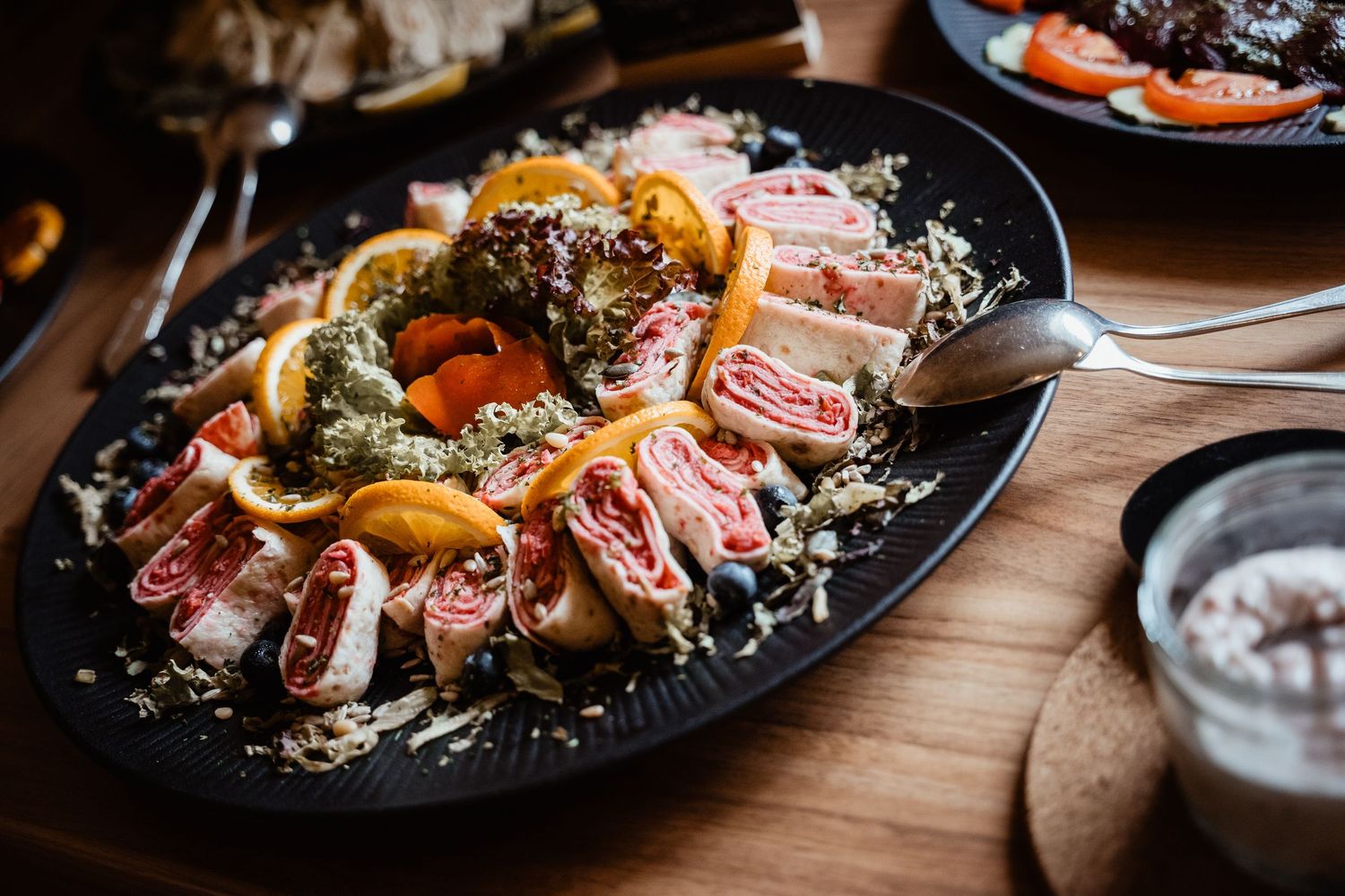 Decorative wedding reception platter with rolled meats, oranges, pesto, and garnishes on dark plate.