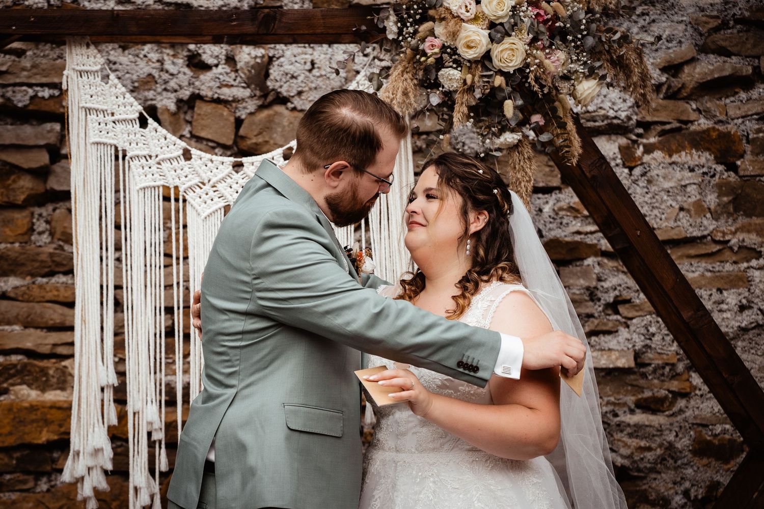 Bride and groom share a tender moment during wedding ceremony before rustic stone wall with macrame backdrop.