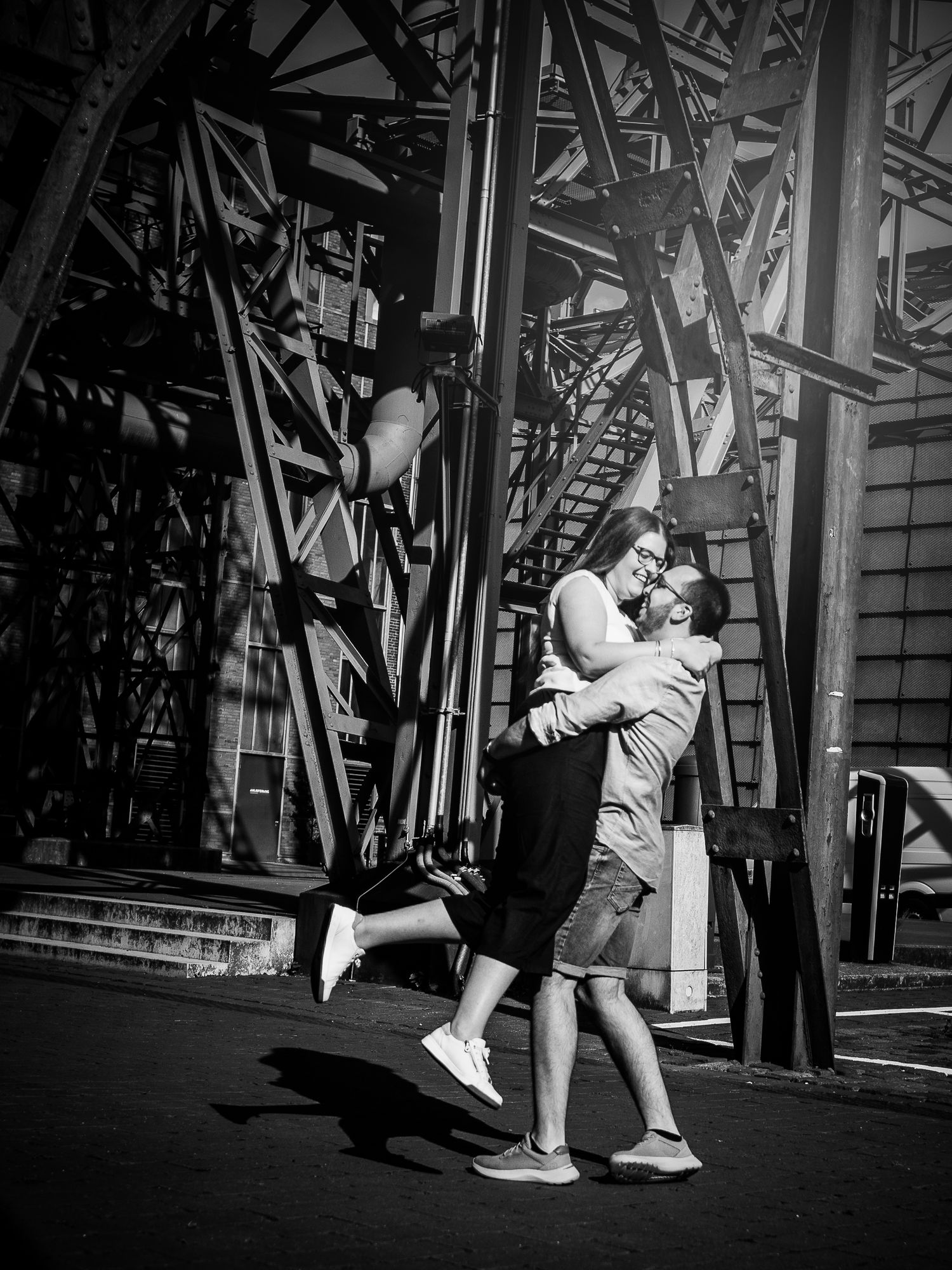 Black and white photo of a couple embracing and kissing near an industrial steel structure outdoors.