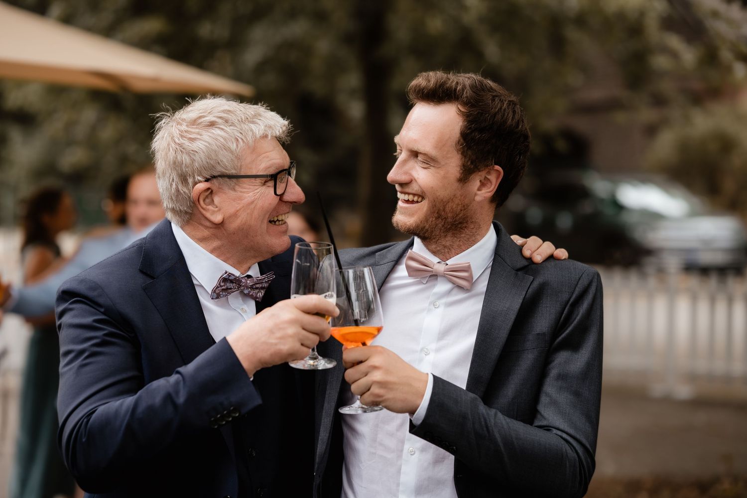 Two men in suits share a cheerful toast with drinks at an outdoor wedding reception.