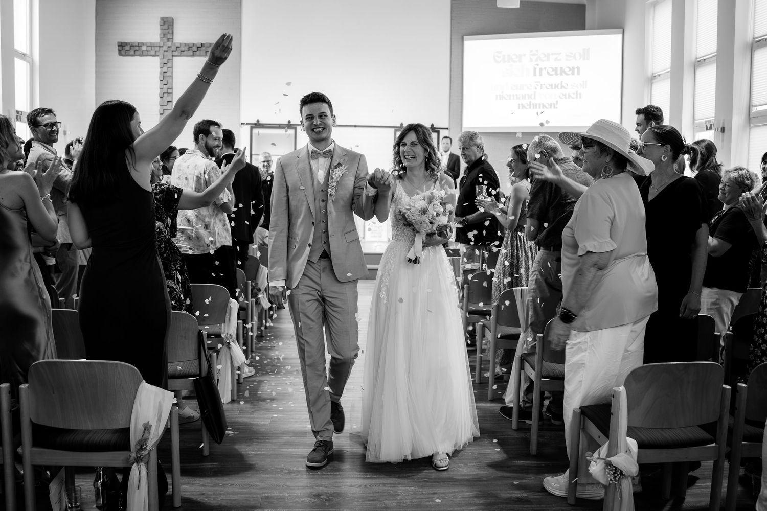 Newlyweds walk down church aisle amid confetti and cheering guests in a joyful black and white wedding photo.