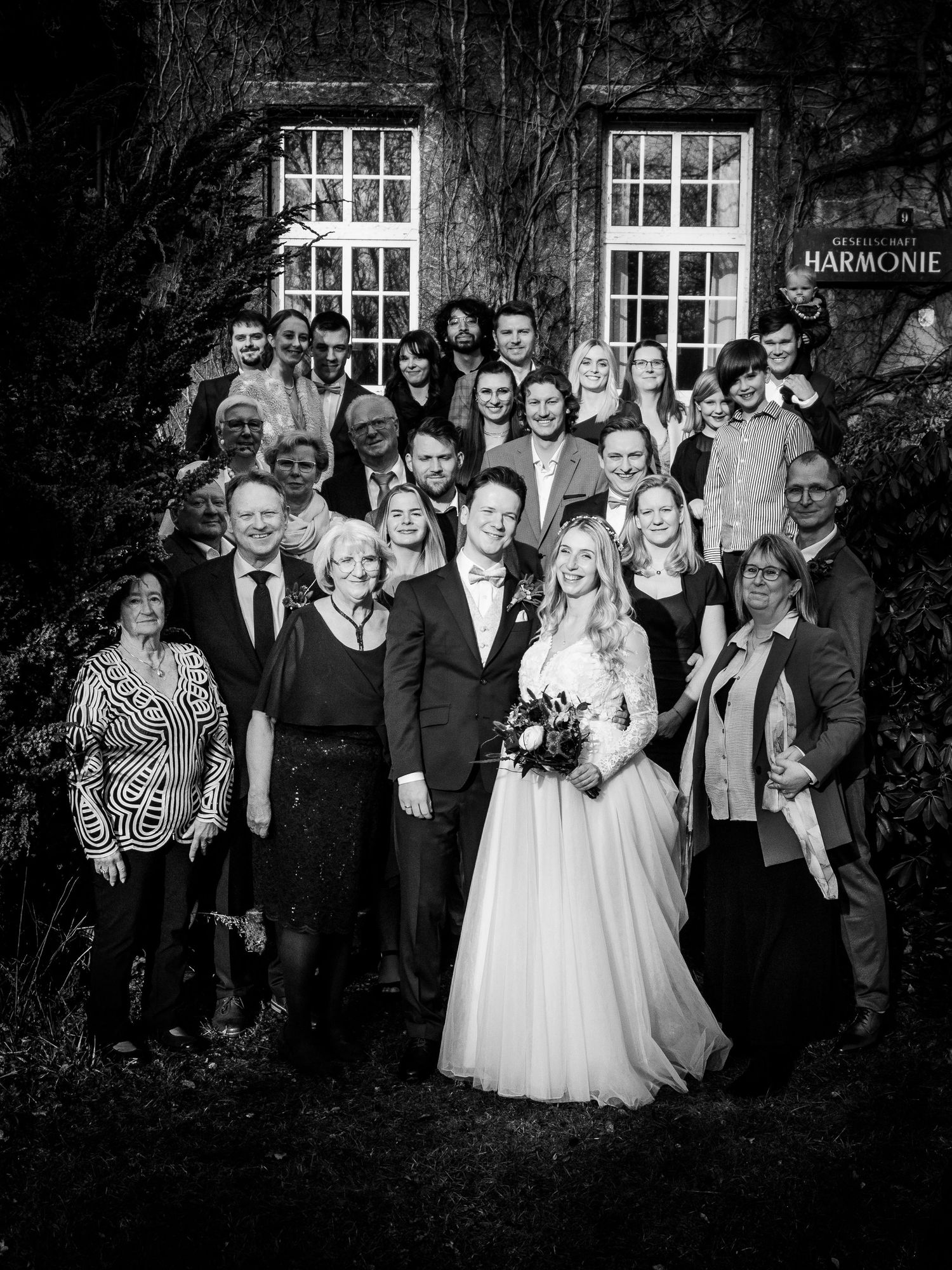 Black and white wedding group photo of couple with guests outside historic Harmonie building.